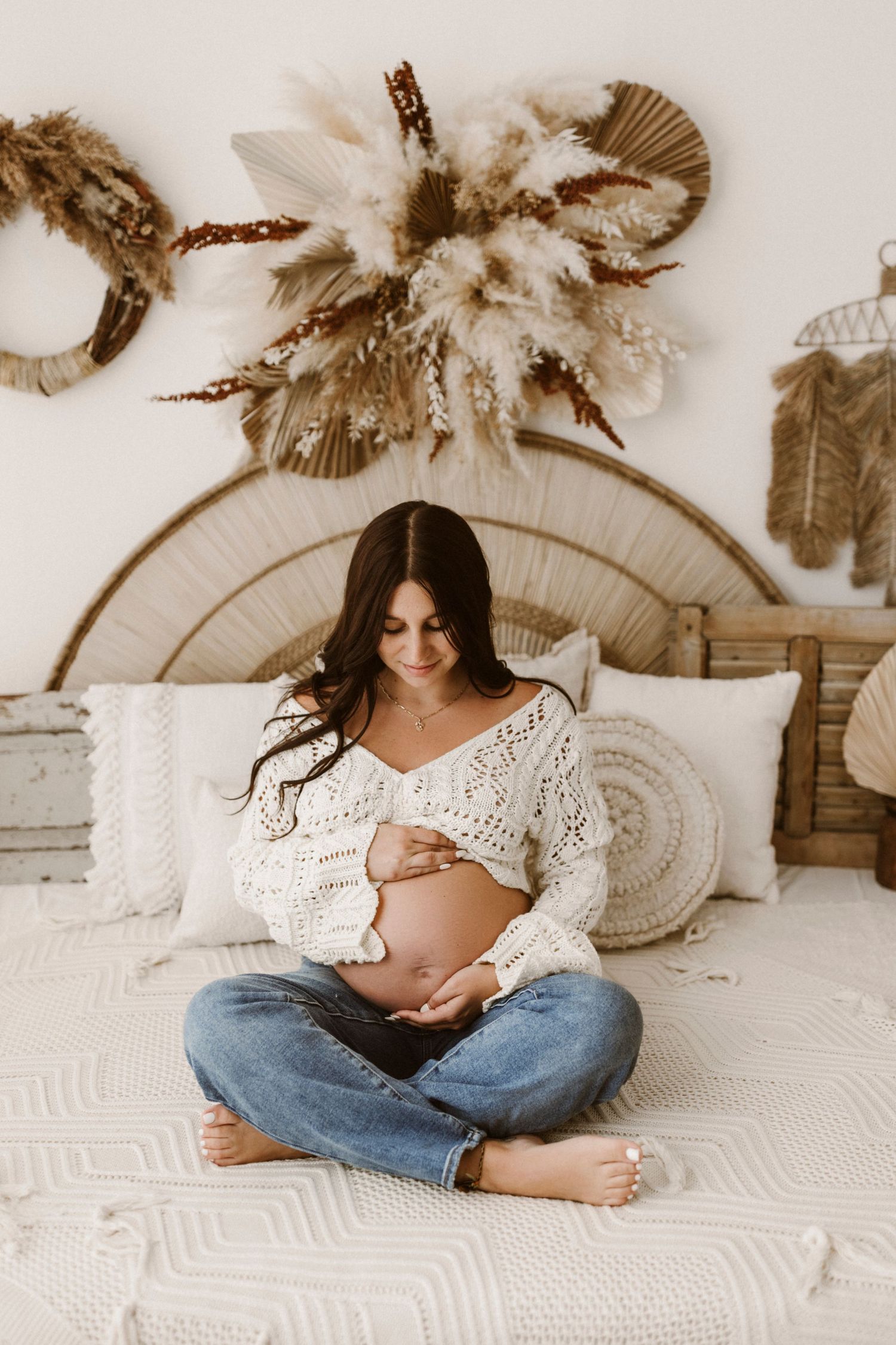 Expecting mother in white sweater and jeans sits cross-legged on bed with boho wall decor and pampas grass above.