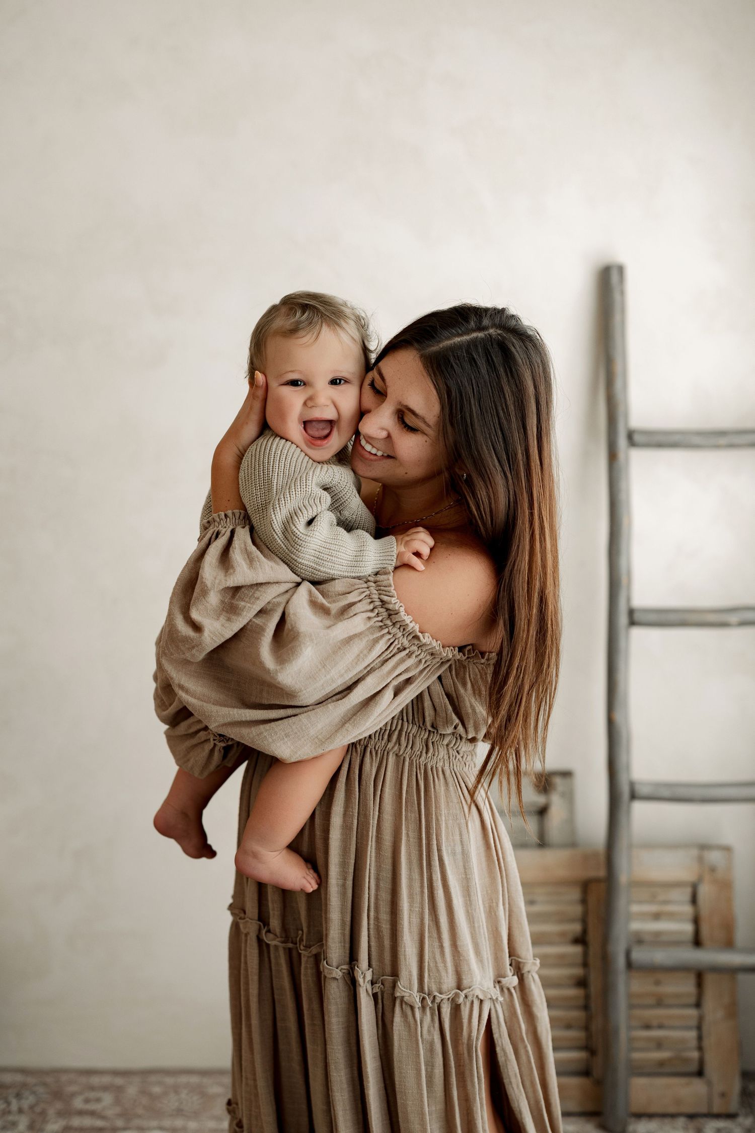 A smiling mother in a beige dress holds her laughing toddler in a bright, minimalist studio setting.