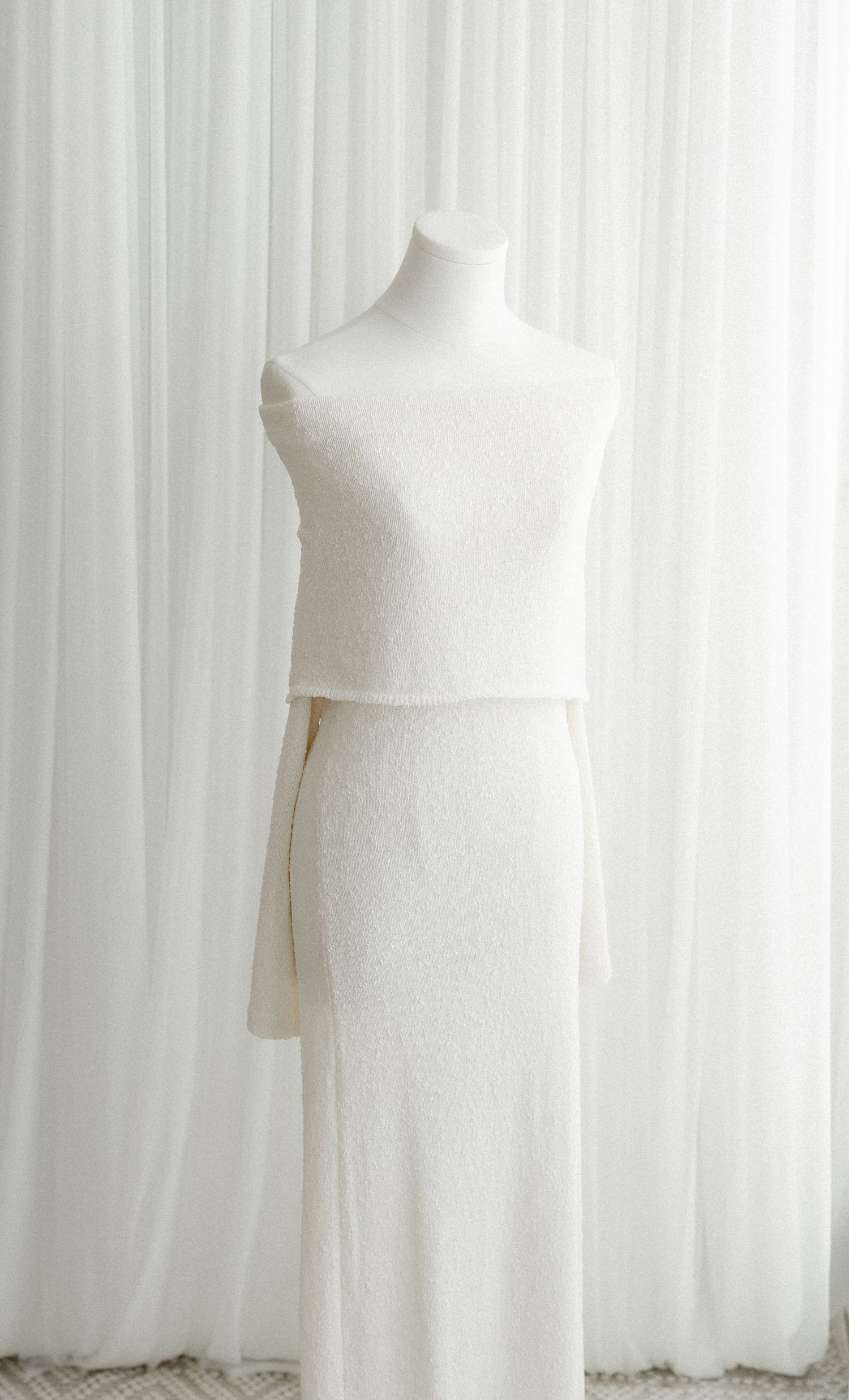 White mannequin dress form stands against sheer white curtain backdrop in minimalist fashion studio setting.