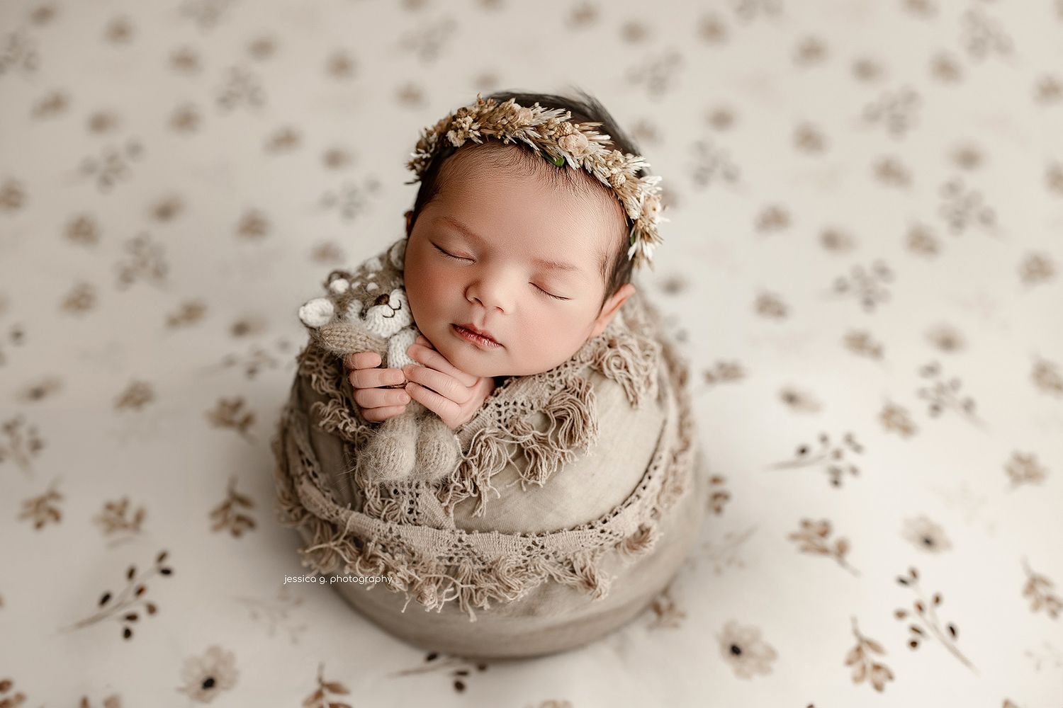 Sleeping newborn wearing a floral crown and beige wrap rests peacefully in a neutral-toned studio setting.