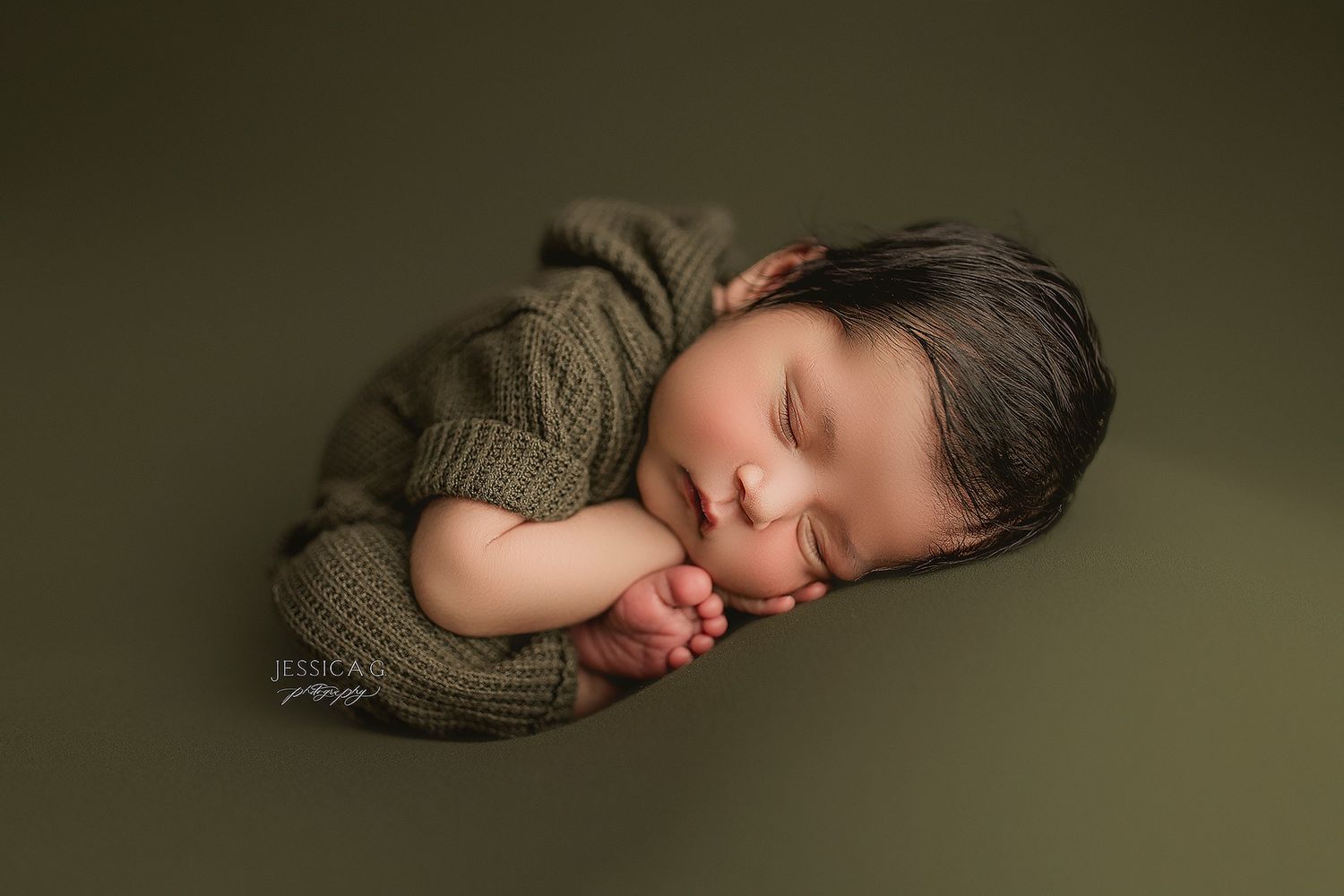 Sleeping newborn baby in olive green backdrop curled up in fetal position wearing neutral colored knits.