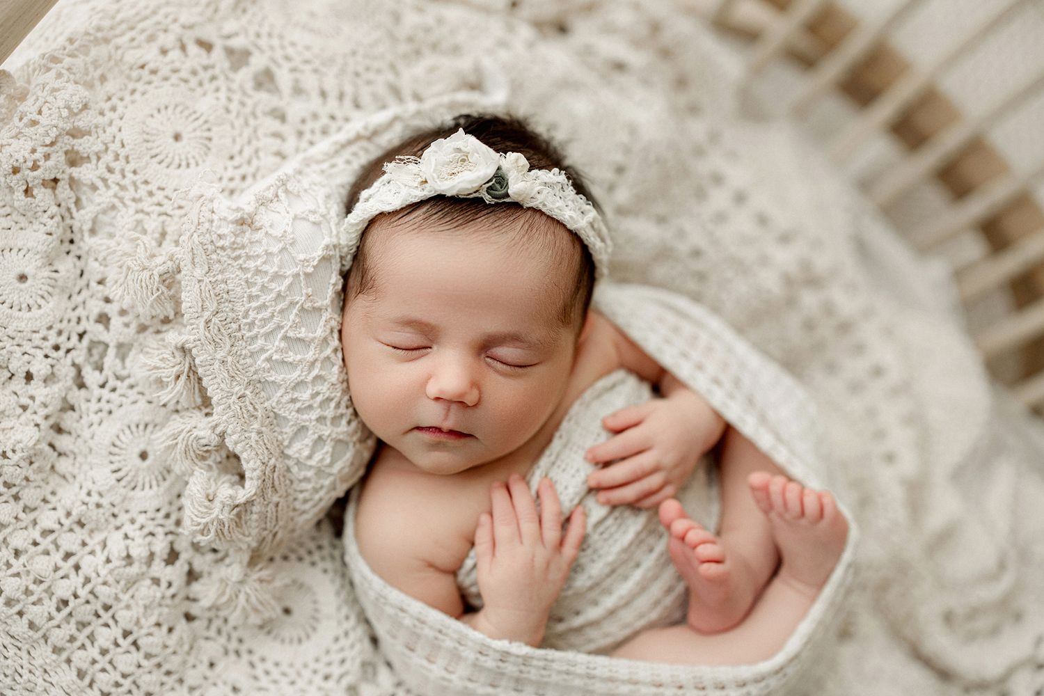 A sleeping newborn rests peacefully in a soft white blanket while lying on textured lace fabric.