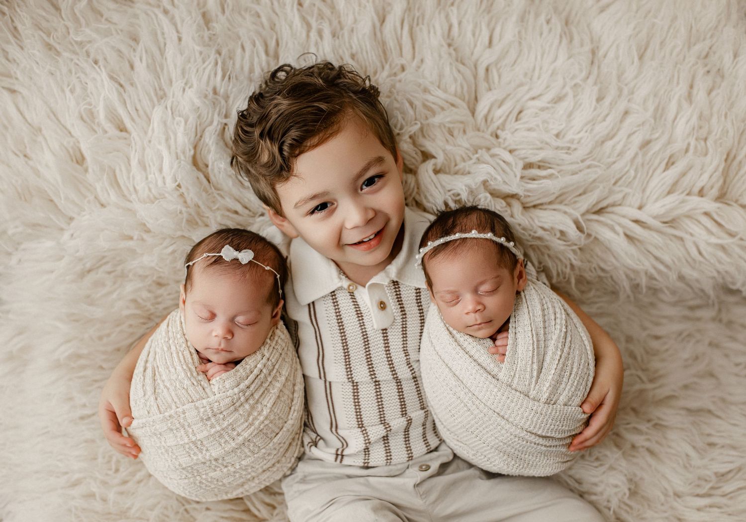 Siblings pose together on soft white fur backdrop as two newborns wrapped in cream blankets snuggle close to their brother.