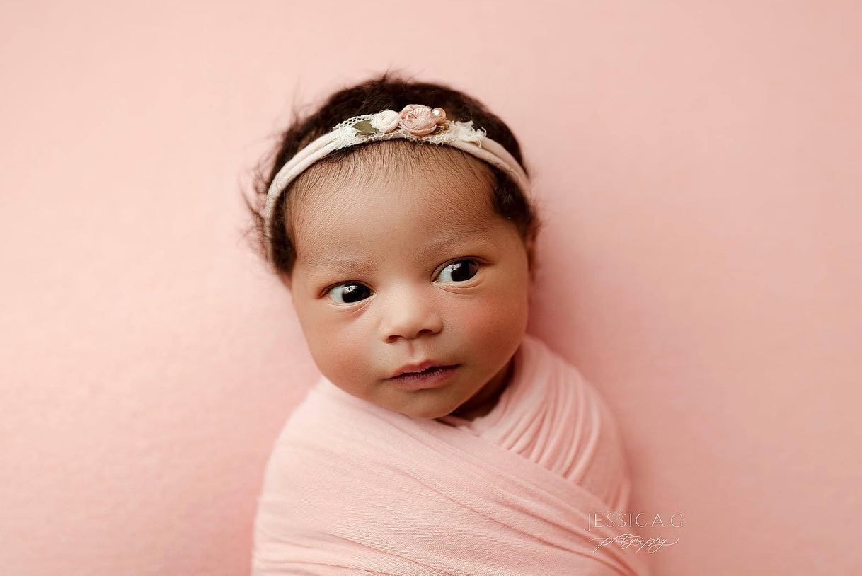 A baby wrapped in soft pink fabric is photographed against a pink background during a newborn photo session.