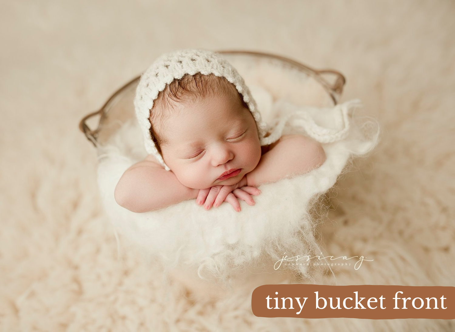 A sleeping newborn baby wearing a white bonnet rests peacefully on cream-colored bedding during a professional photo shoot.