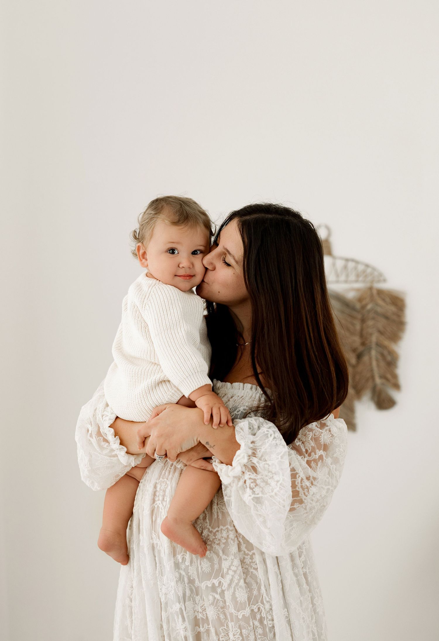 A mother in a white lace dress kisses her baby girl dressed in white, against a soft neutral background.
