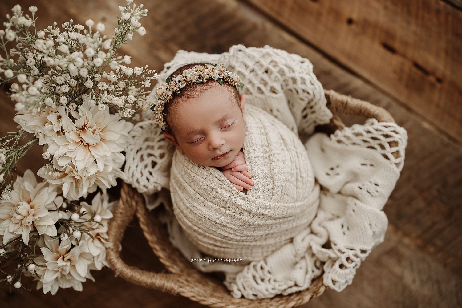 Sleeping newborn wrapped in cream knit blanket nestled in basket with white floral decorations on wooden floor.