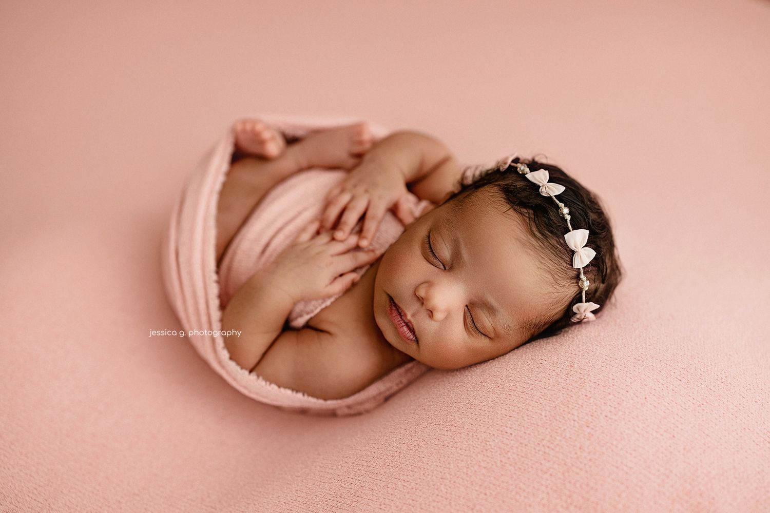 Newborn baby wrapped in pink blanket wearing delicate flower crown sleeps peacefully on pink background.