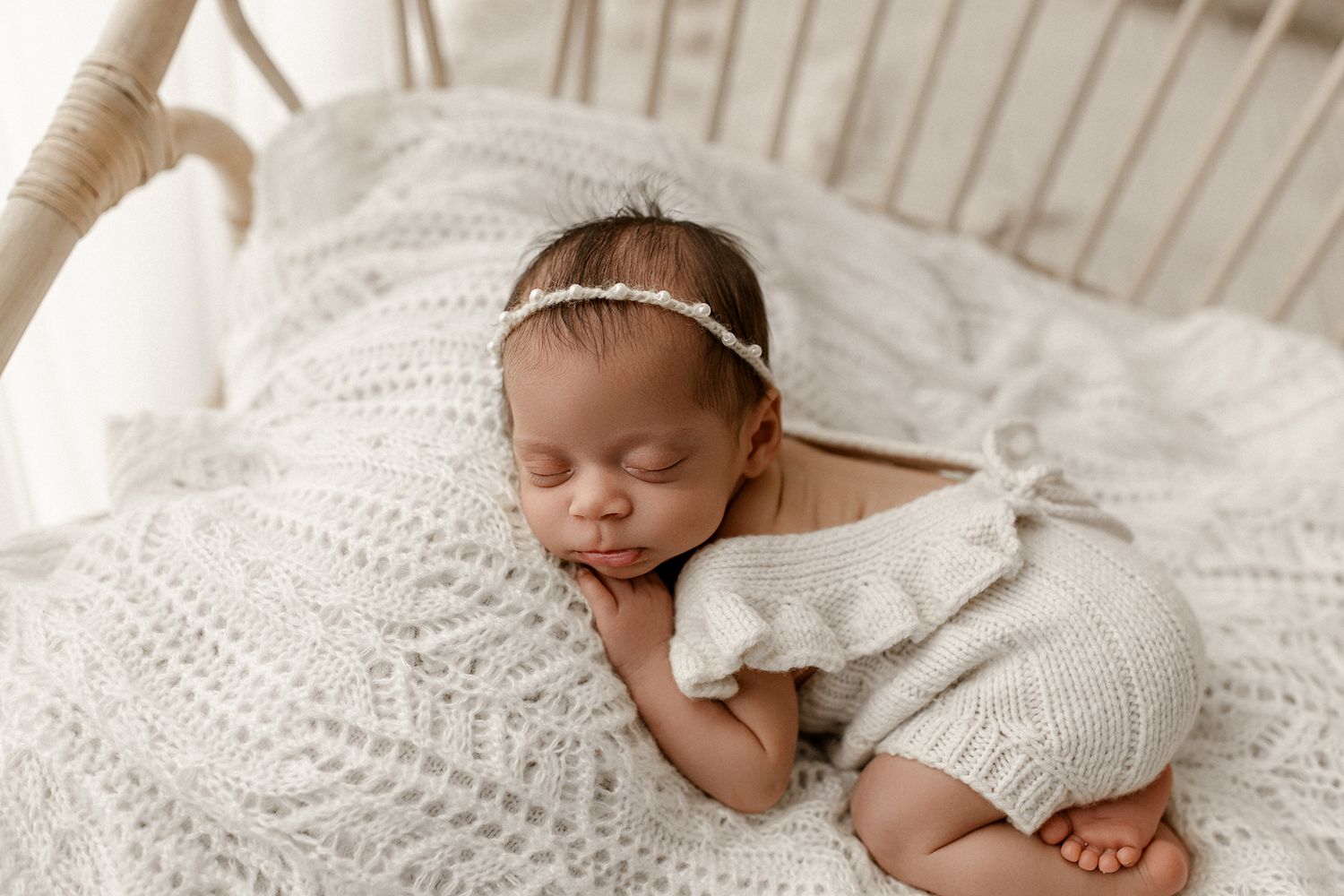 A sleeping newborn baby wearing a white knit outfit rests peacefully on textured cream bedding.