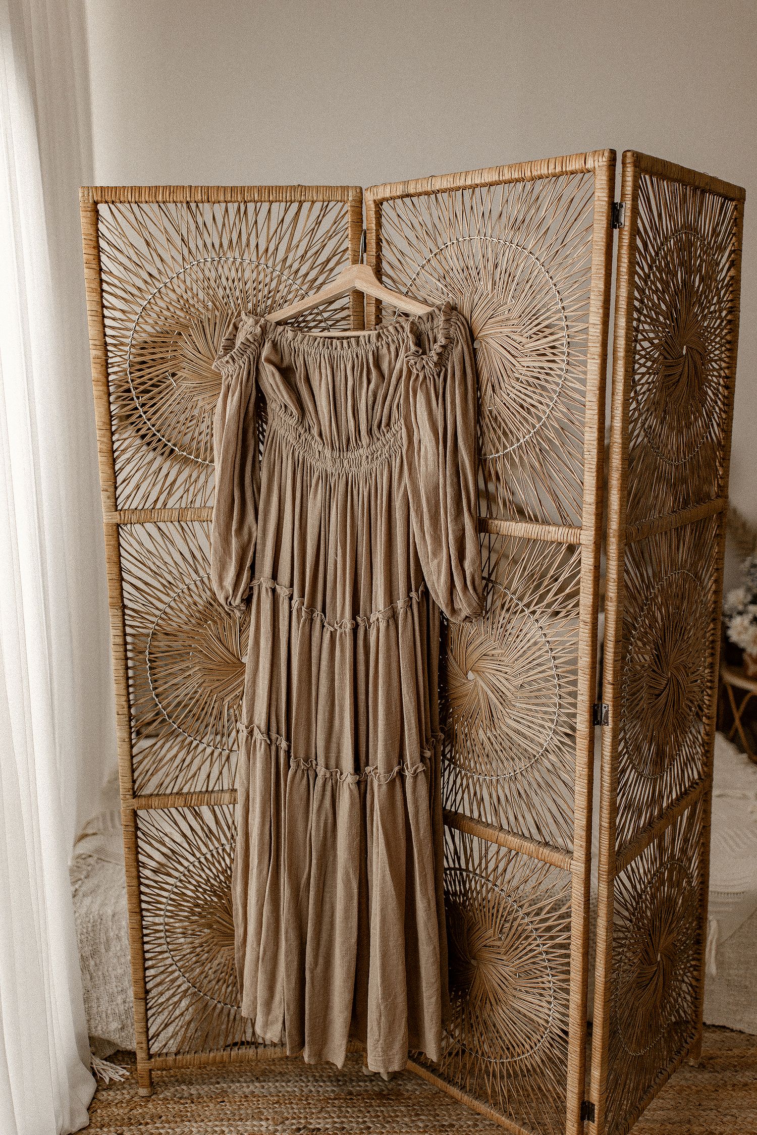 A beige tiered maxi dress hangs on a woven rattan room divider with sunburst pattern panels.