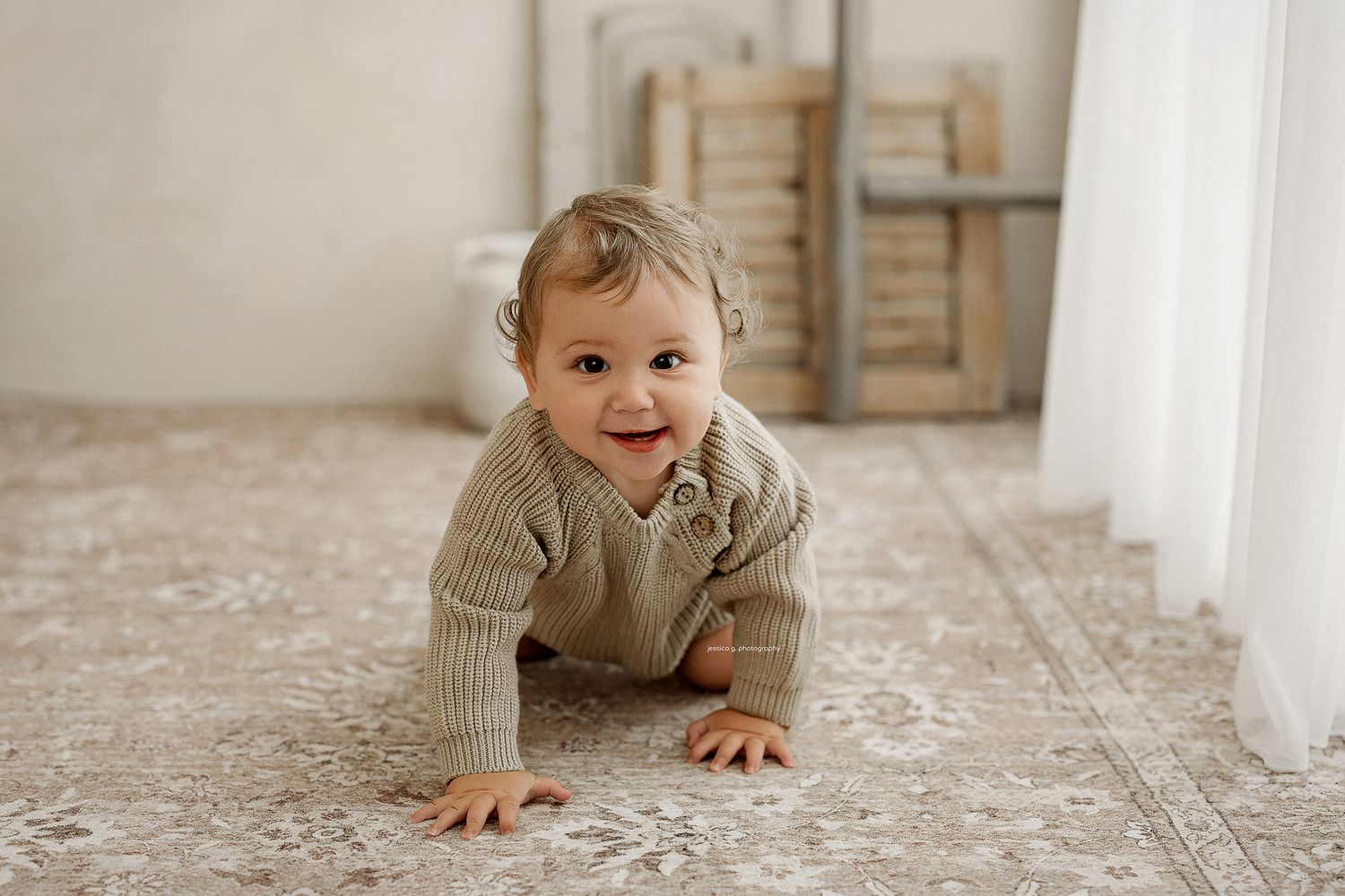 A smiling baby in a knit romper crawls on a patterned rug in a bright, minimalist room.
