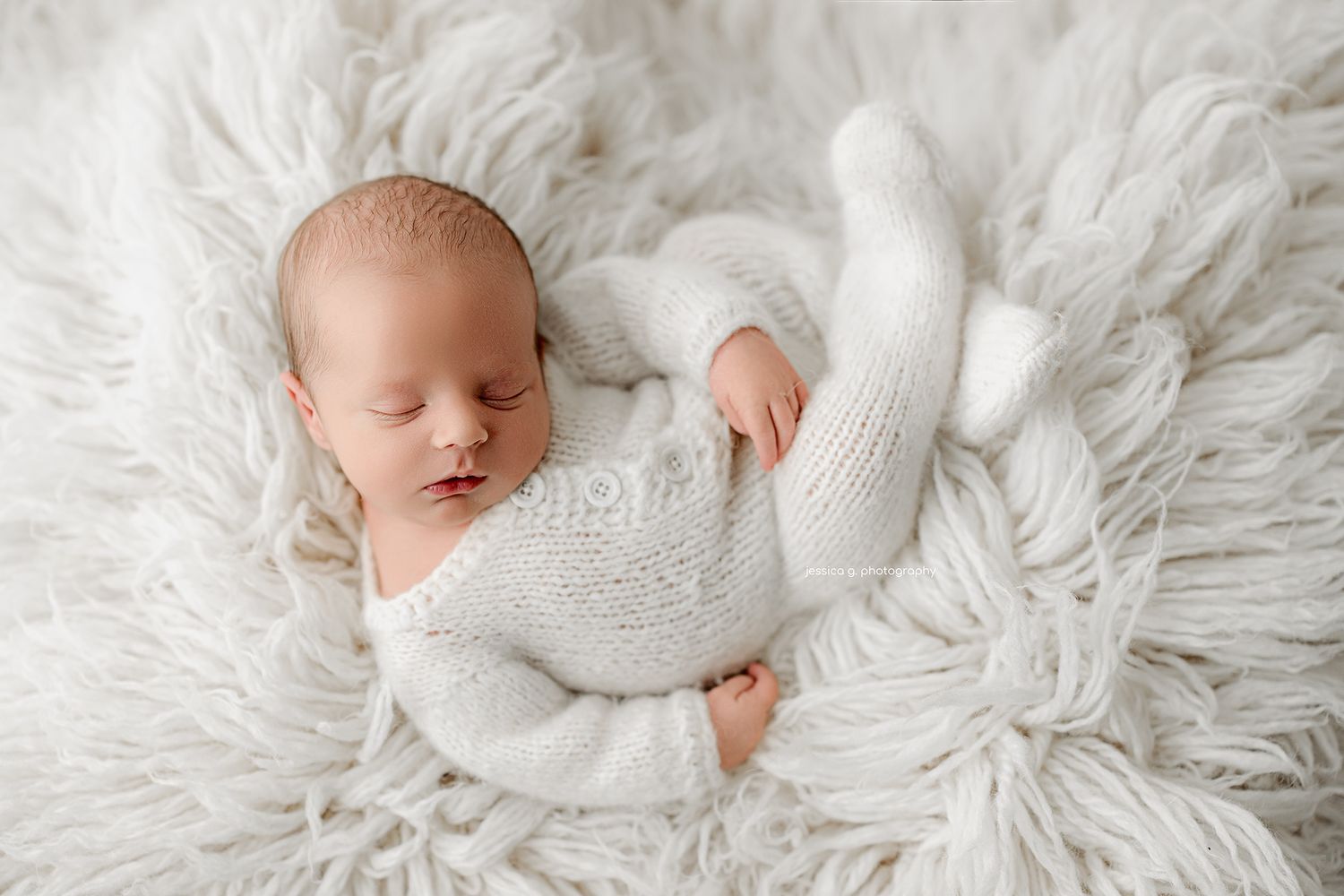 A sleeping newborn in a white knit outfit lies peacefully on soft white fur blanket.