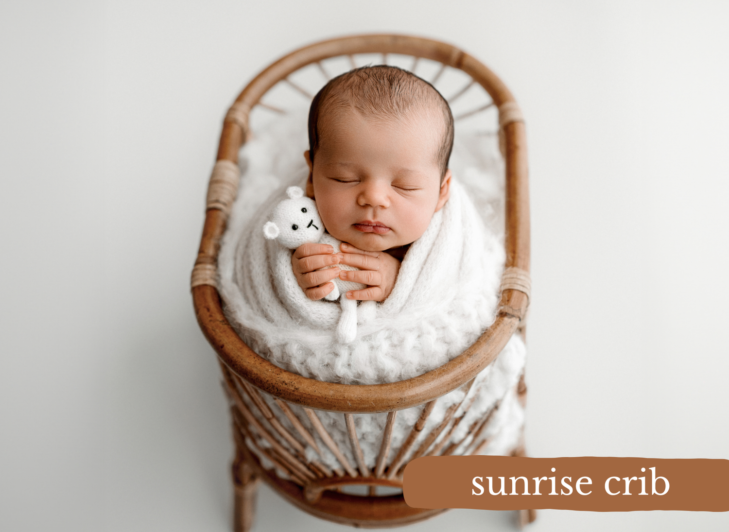 A newborn rests peacefully in a wooden basket lined with soft white fabric, photographed in warm natural light.