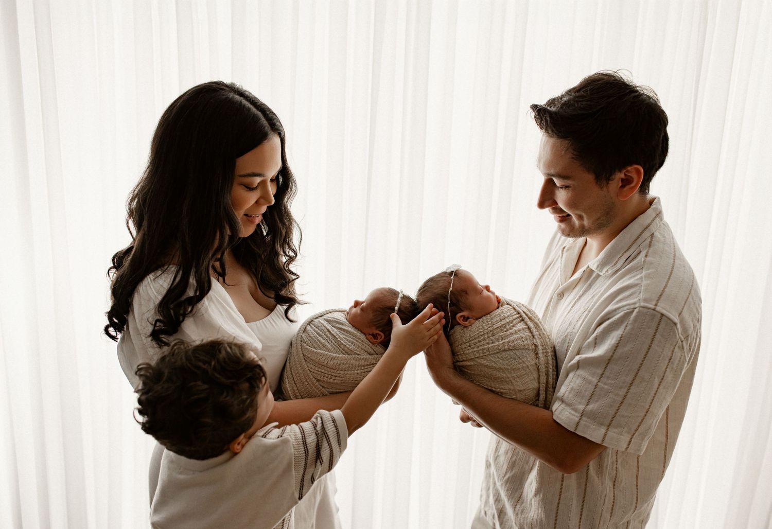 New parents cradling their sleeping baby together while standing in front of white curtains in natural light.