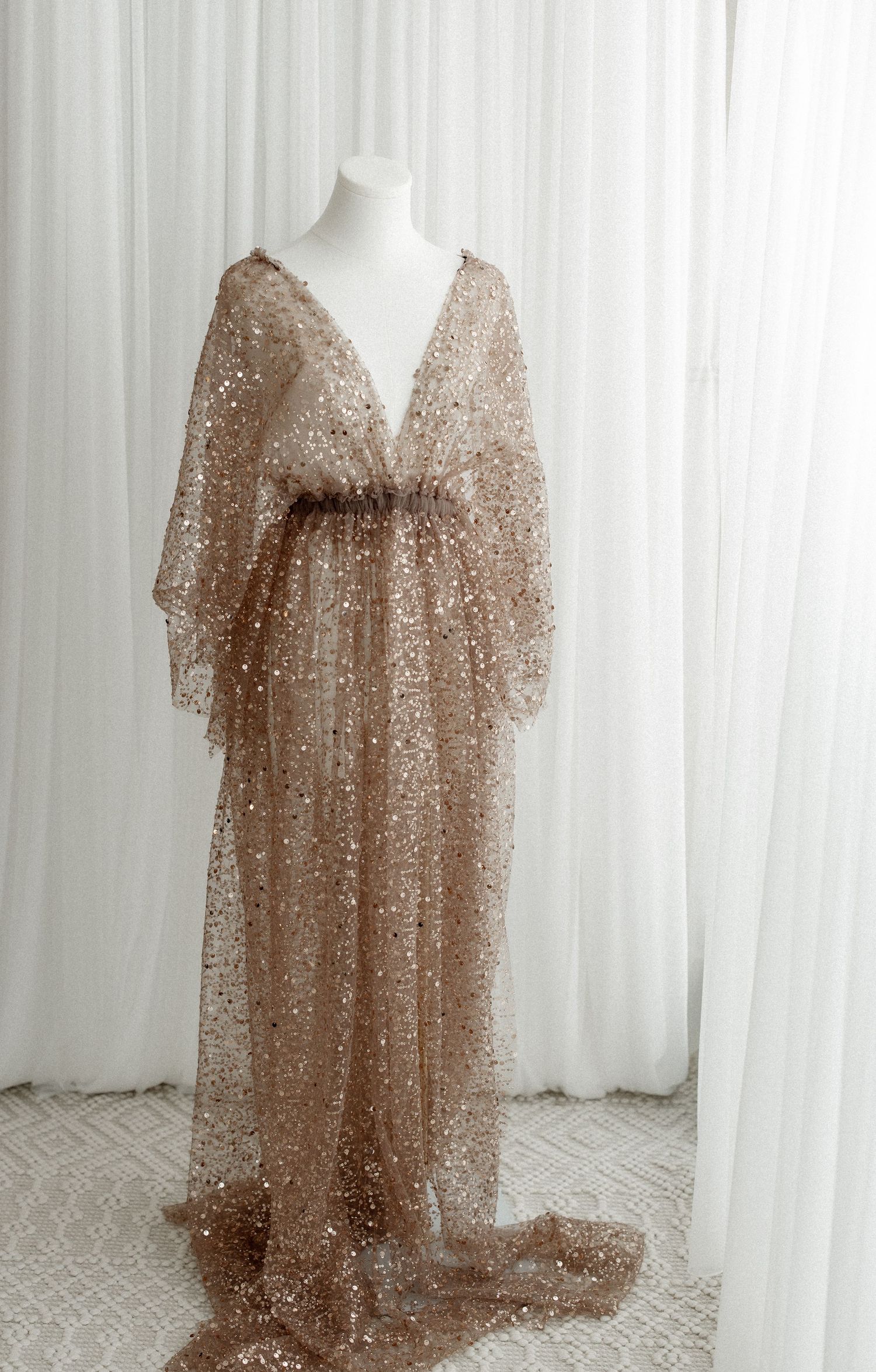 Elegant rose gold sequined evening gown with flowing batwing sleeves and plunging V-neckline on mannequin display.