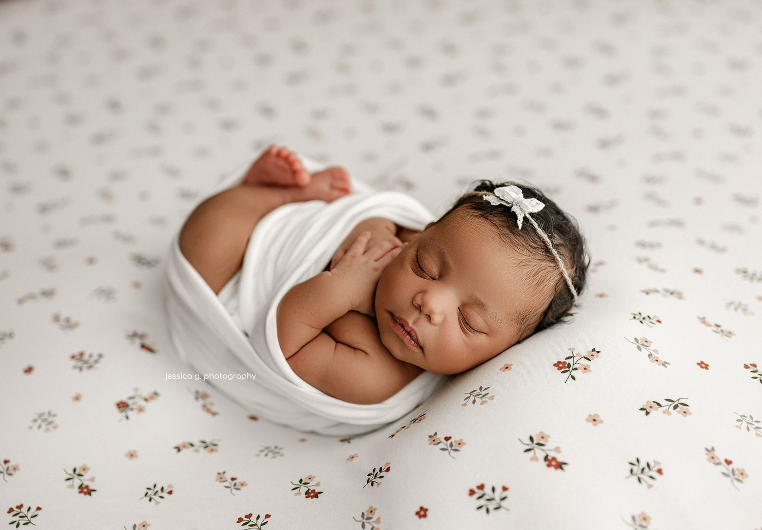 Sleeping newborn wrapped in white swaddle blanket rests on floral patterned fabric wearing delicate headband.