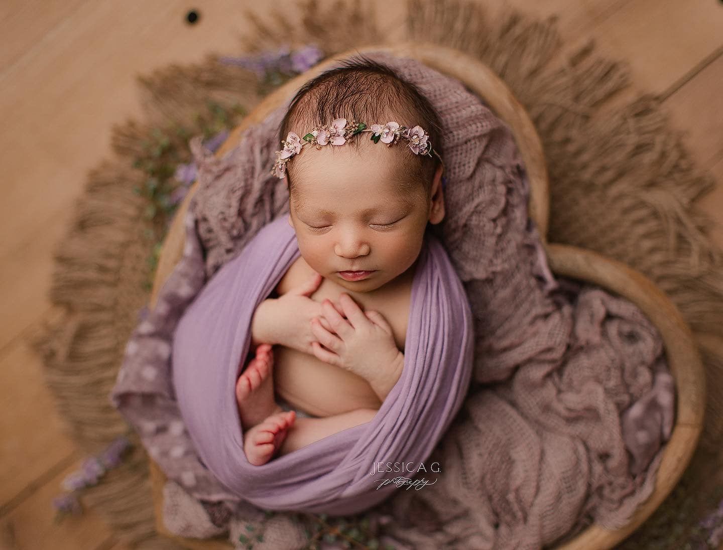 Sleeping newborn wrapped in soft purple fabric nestled in woven basket with delicate floral headband.