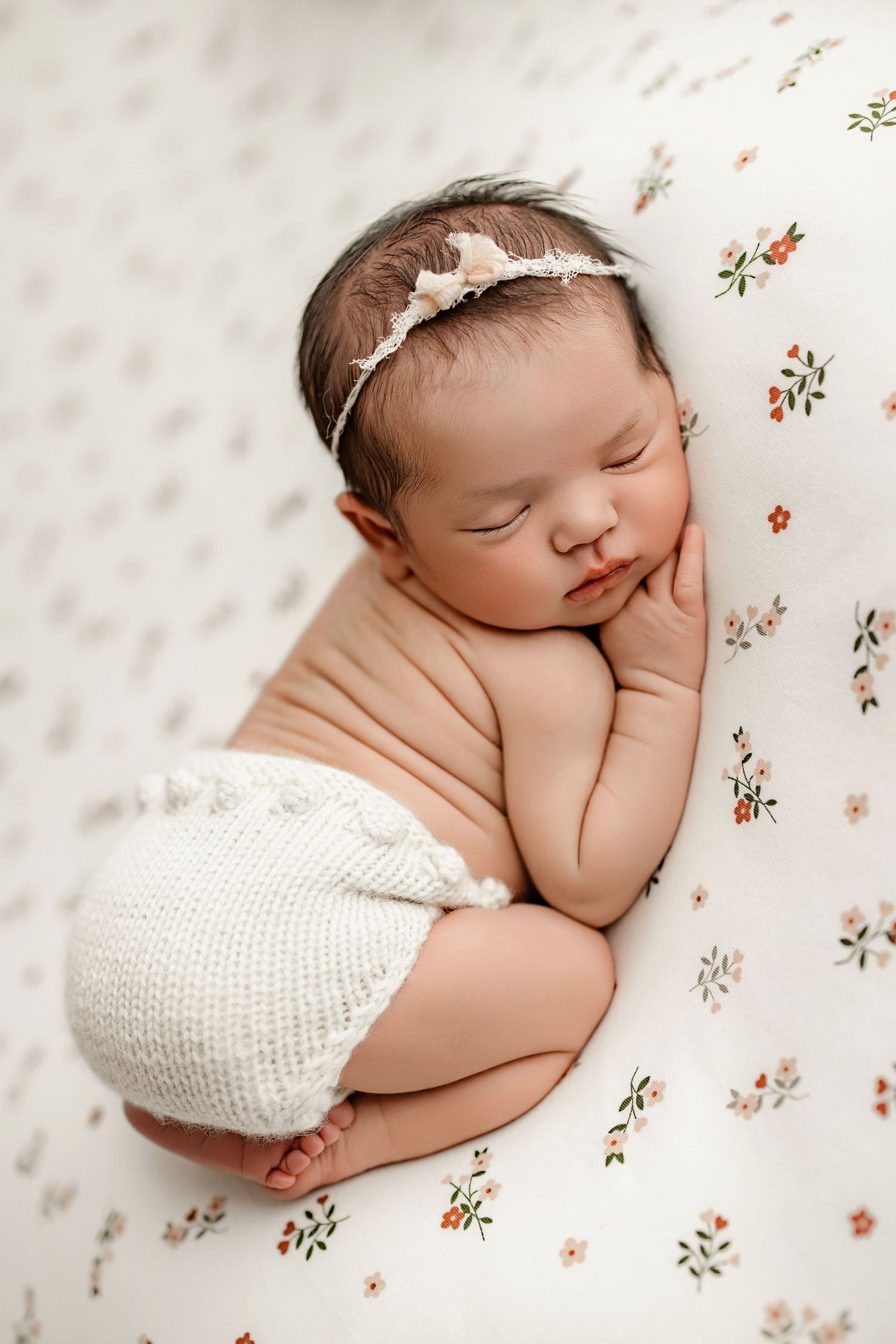 Sleeping newborn in white knitted outfit on floral fabric with delicate headband.