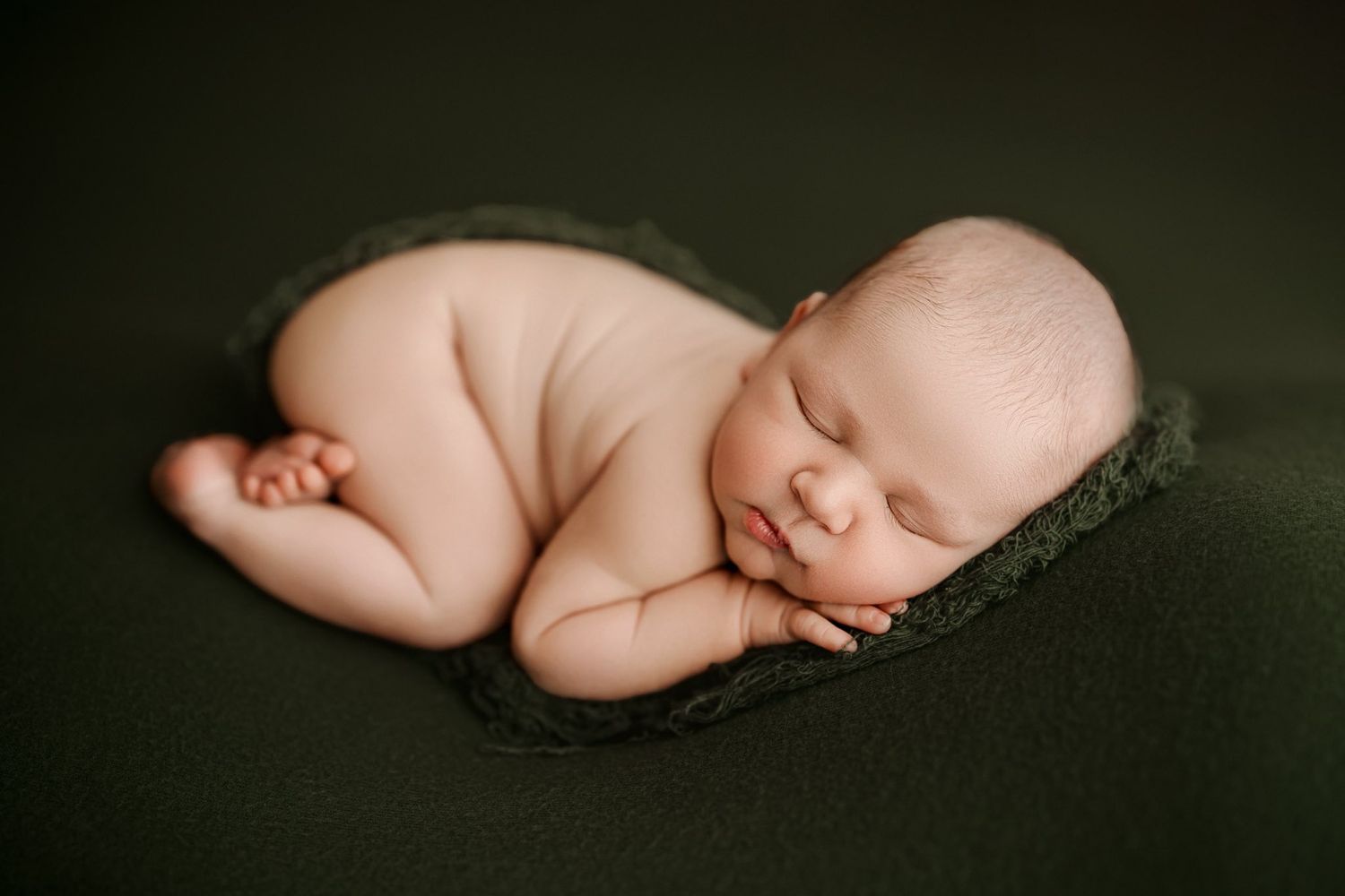 A sleeping newborn baby lies peacefully curled up on a dark background in a minimalist portrait.