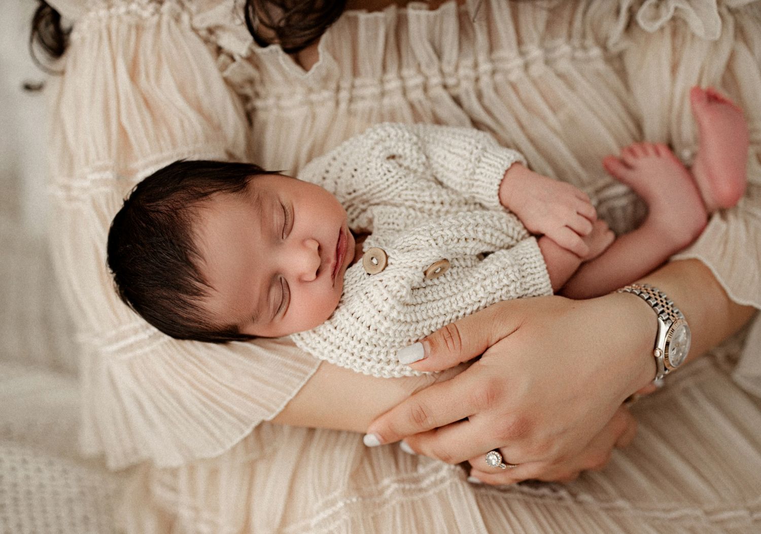 Newborn baby sleeping peacefully in cream knit outfit, cradled in mother's hands wearing a silver bracelet.