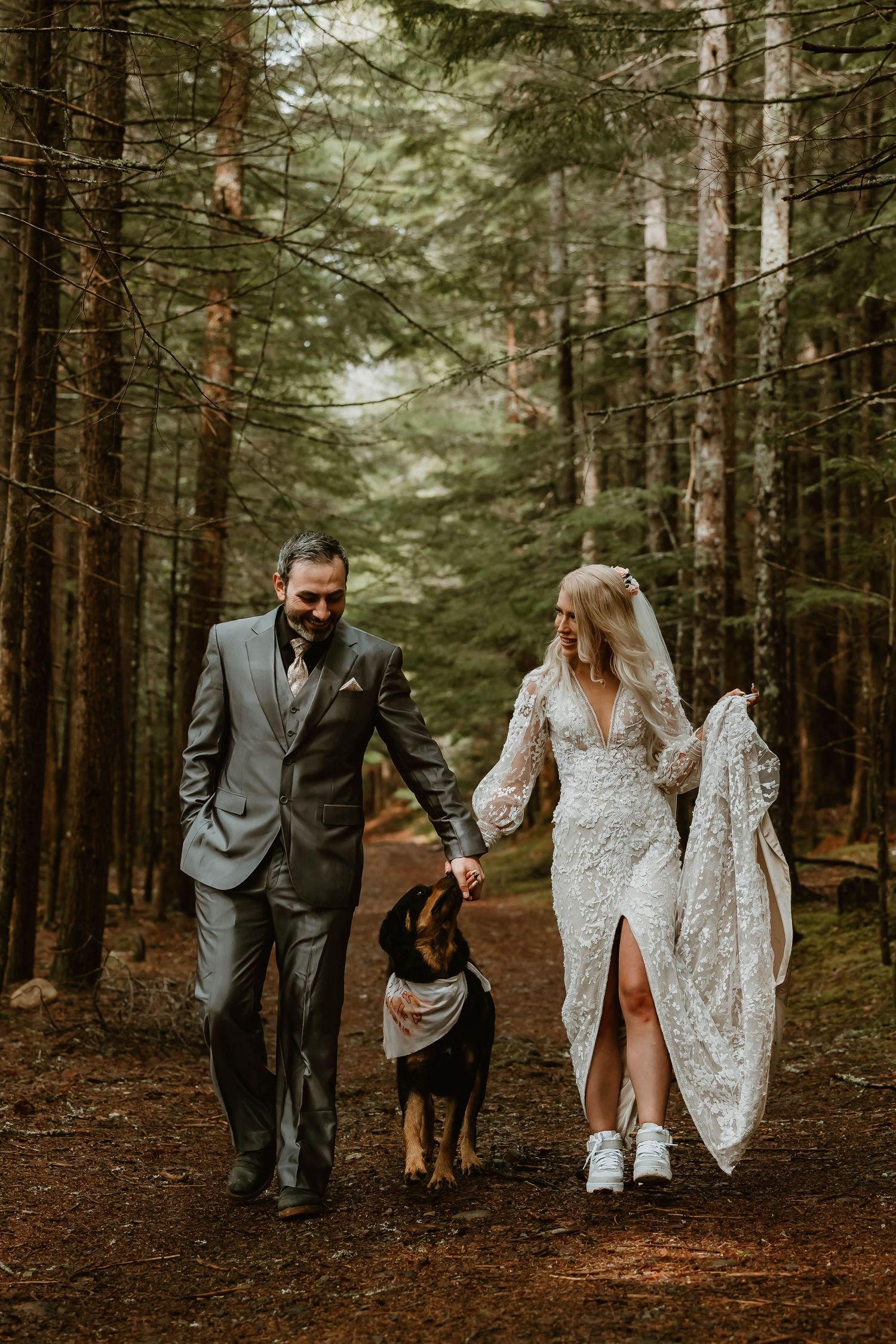 A couple in wedding attire walks through a forest path with their dog, surrounded by tall evergreen trees.