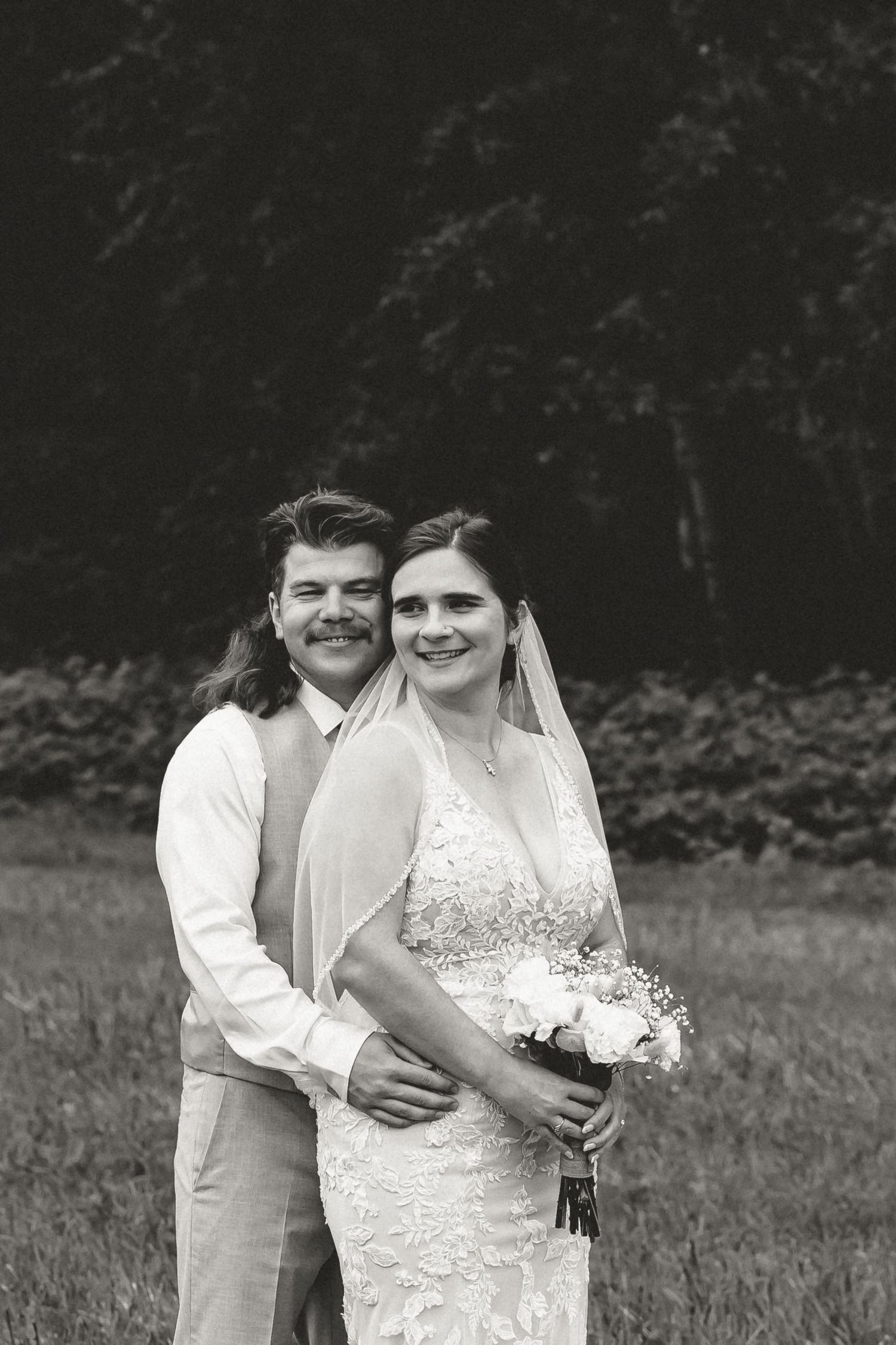 Black and white wedding photo showing a romantic moment between a couple in a field with trees in the background.