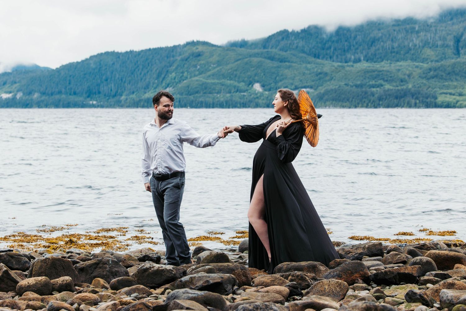 A couple dances on rocky shoreline with mountains and water in background.