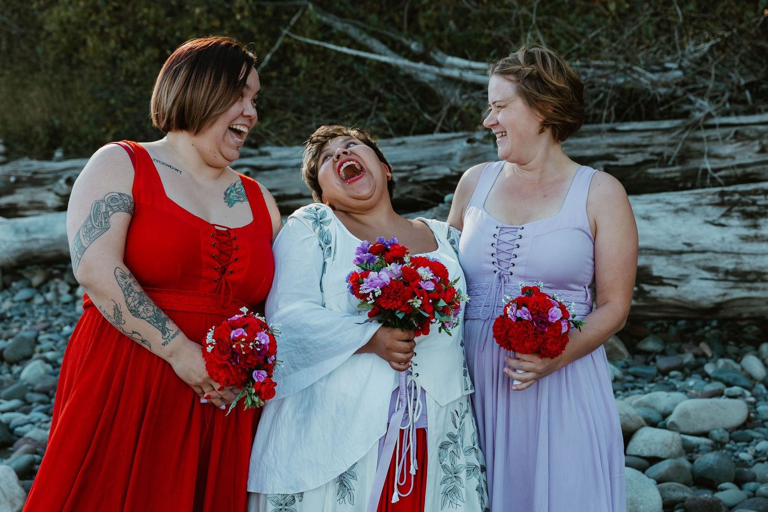 Bridal party laughing together while holding red bouquets outdoors on rocky terrain.