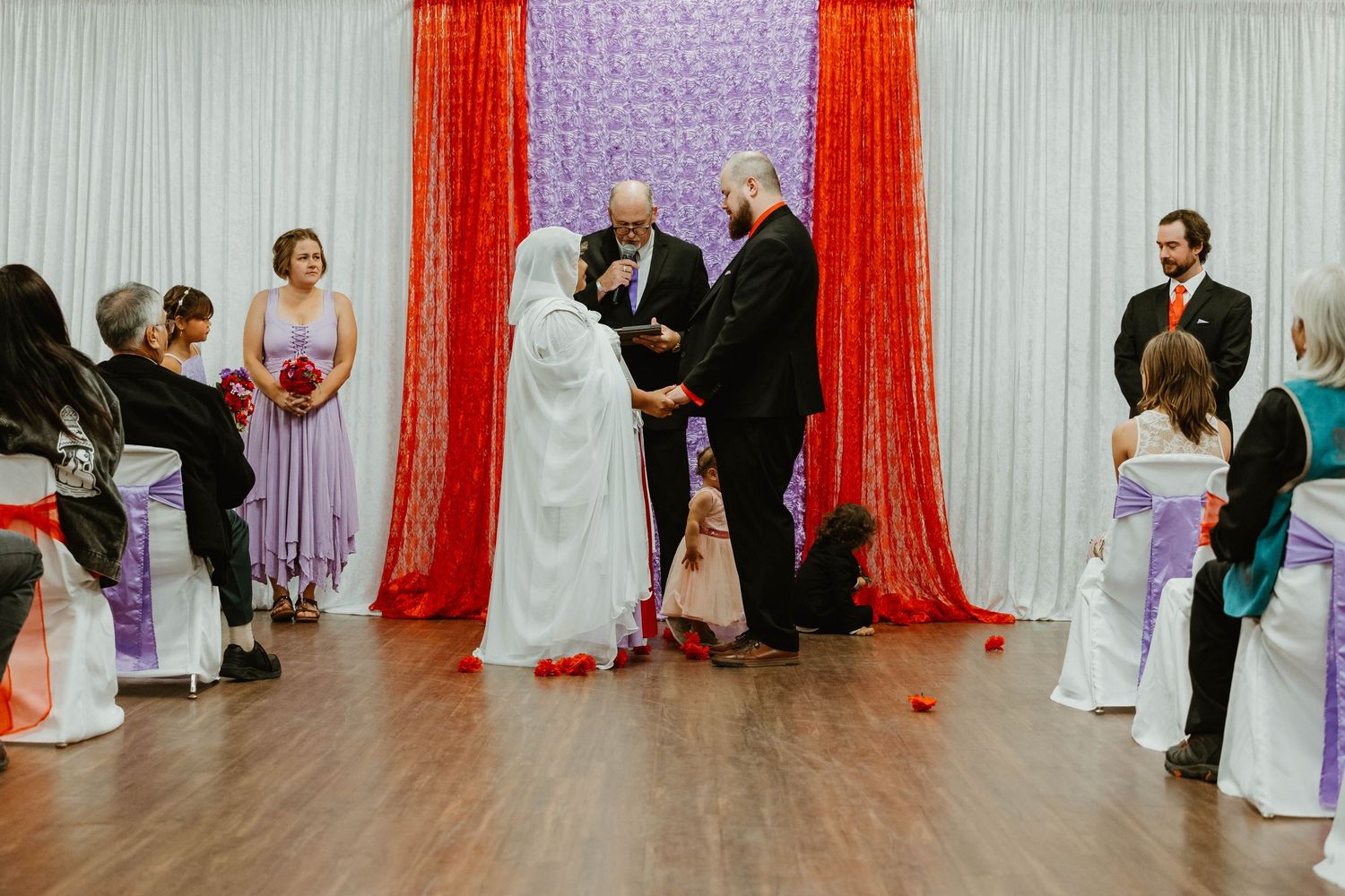 Wedding ceremony in progress with red and white drapery decorations and guests seated on white chairs.