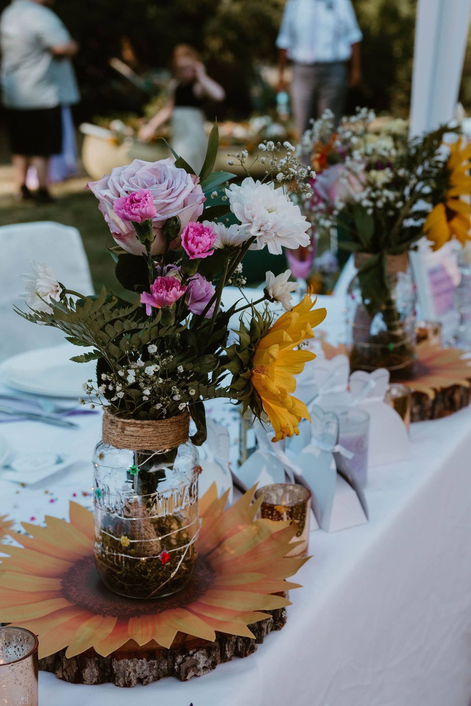 Mason jar centerpiece with pink roses and sunflowers on wooden slice at wedding reception.