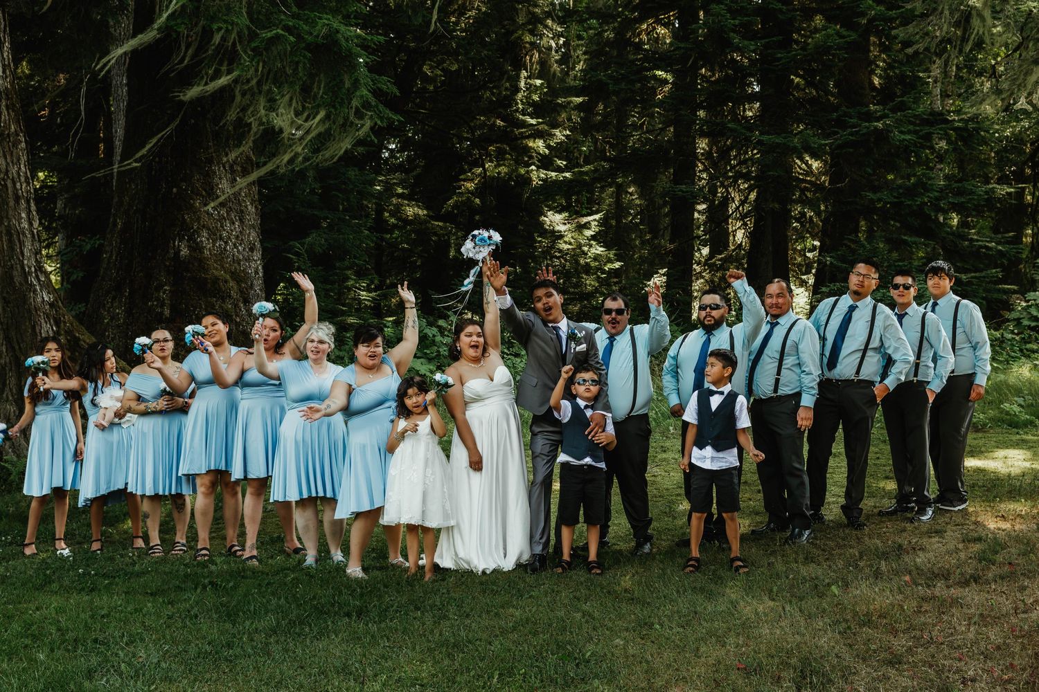 Wedding party in light blue attire celebrating outdoors with raised hands.