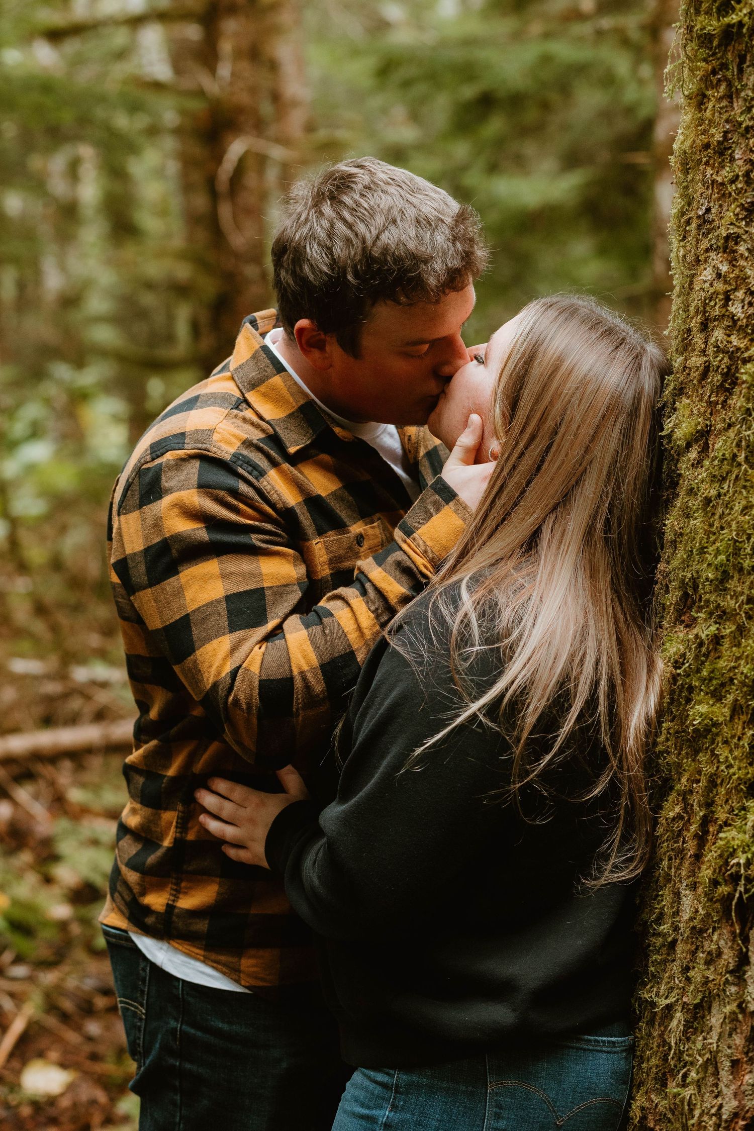 A couple sharing an intimate moment against a moss-covered tree in a forest.