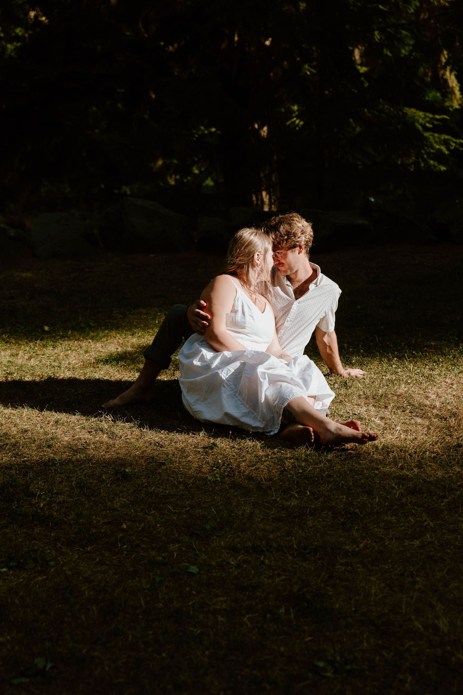 A romantic couple sits together in dappled sunlight on grass wearing white summer clothing.