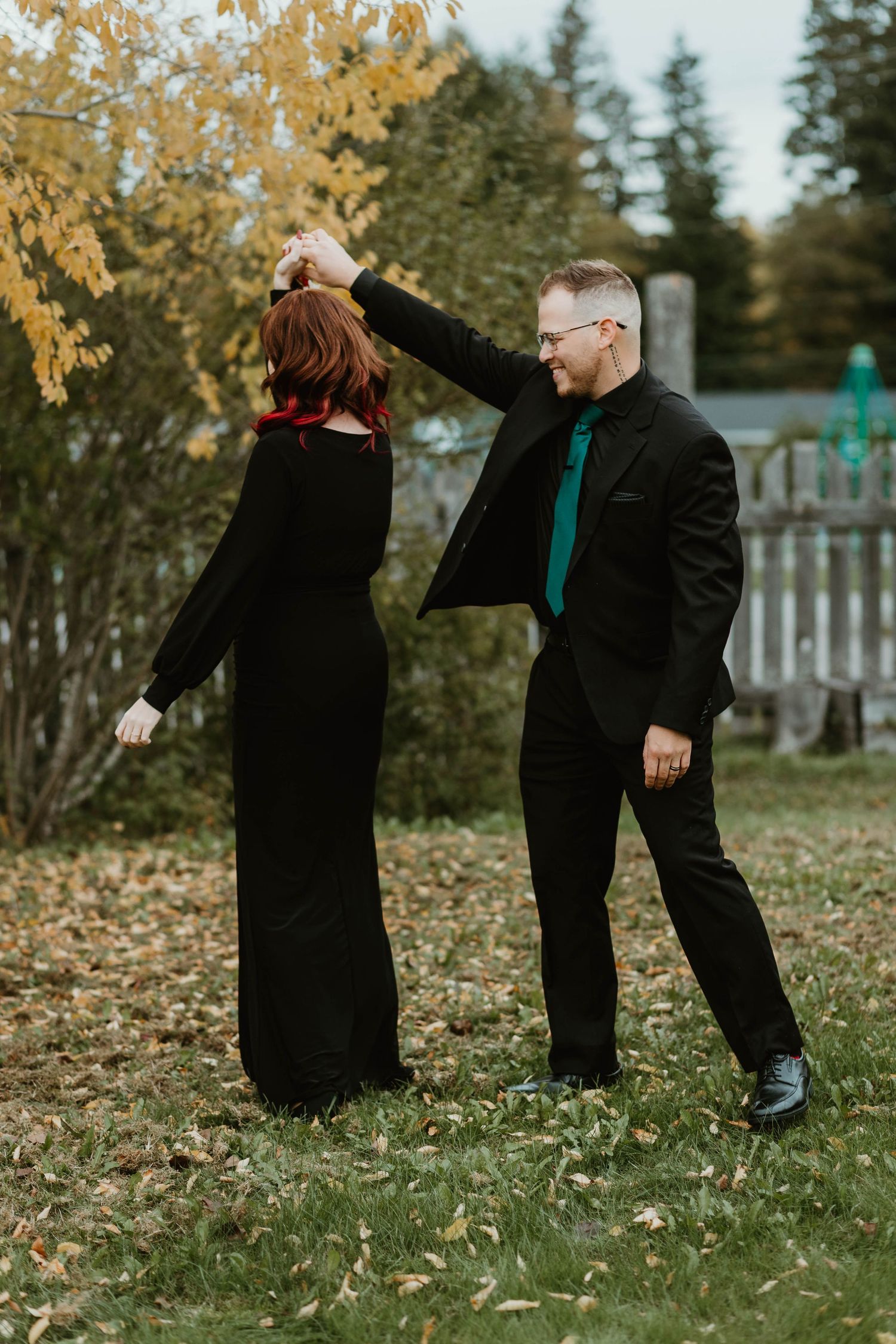 A couple in formal black attire dancing outdoors among fall foliage.