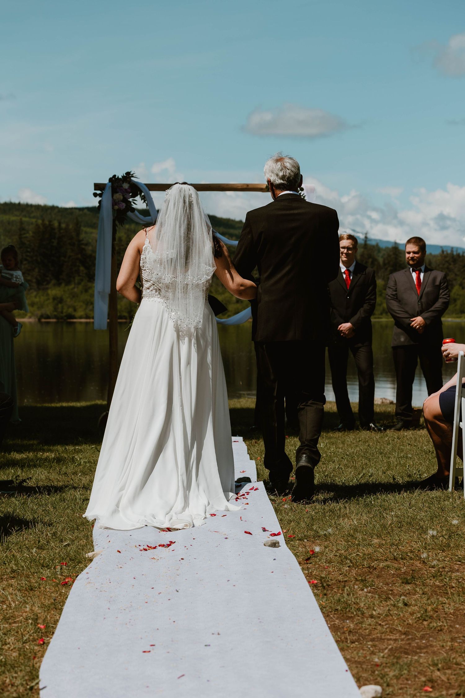Bride in white dress walks down outdoor aisle with escort against lakeside mountain backdrop.