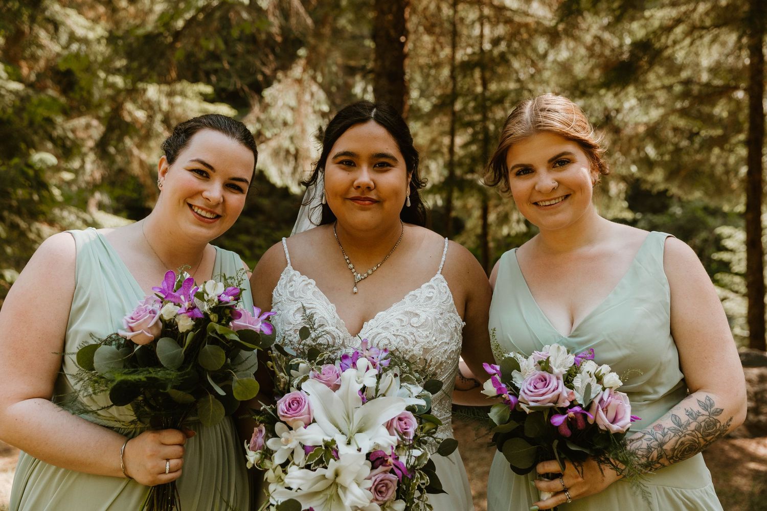 Wedding party members pose with purple and white floral arrangements in outdoor setting.