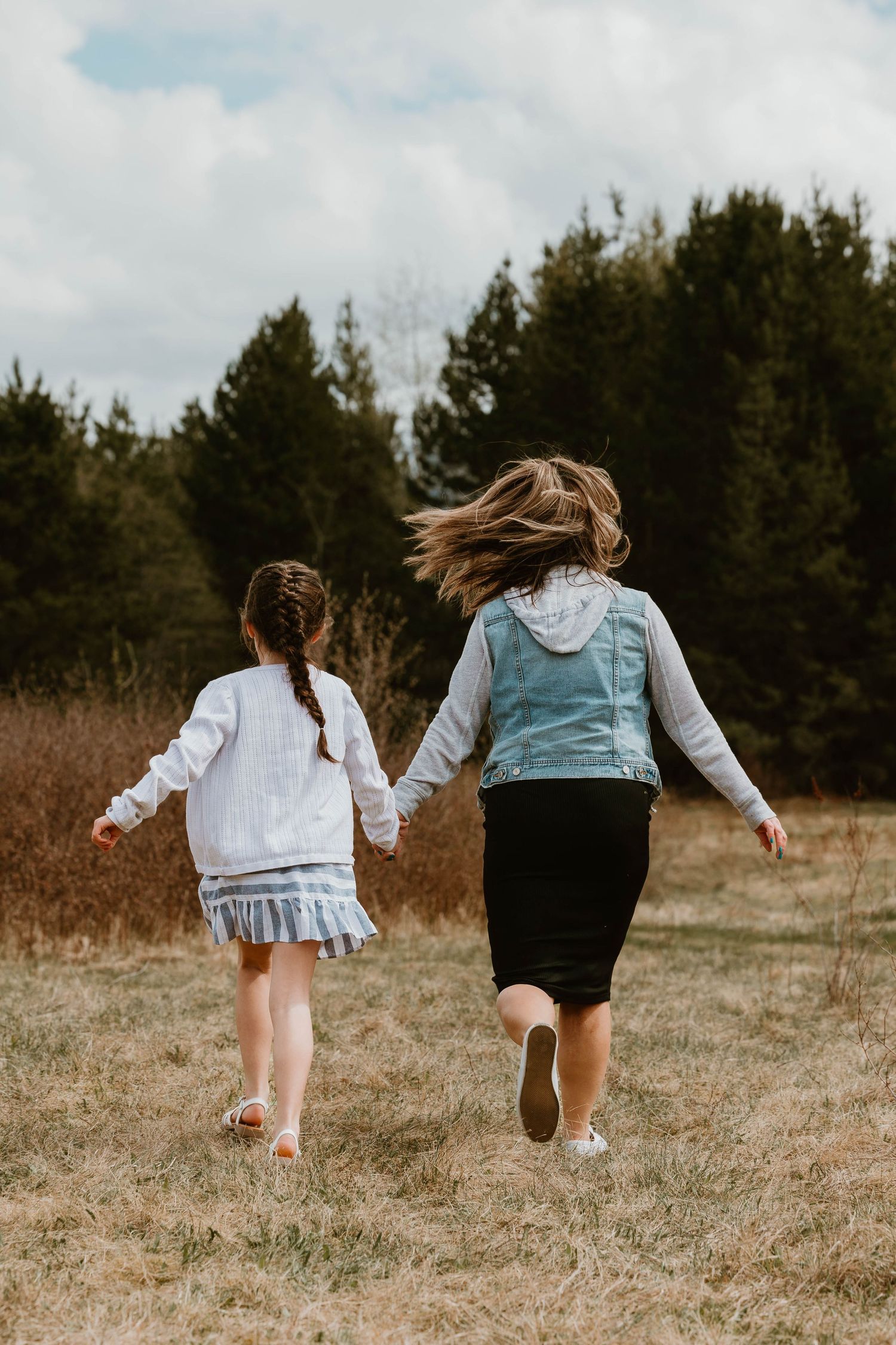 Two people run together through a grassy field with evergreen trees in background.