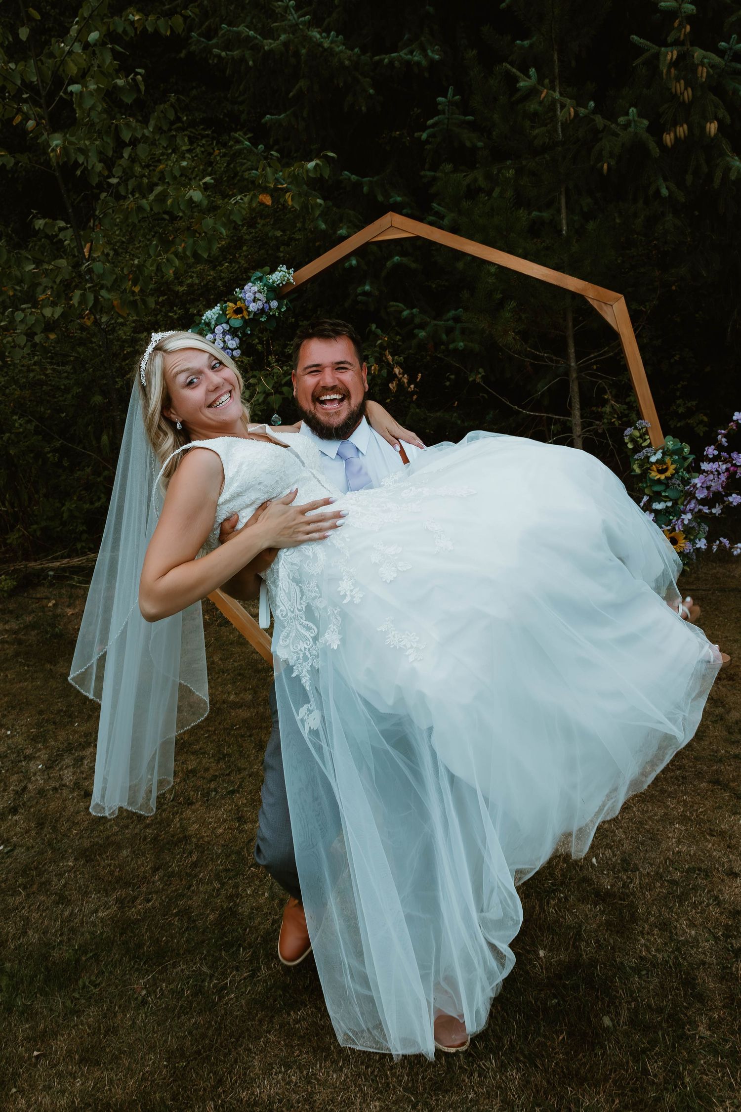 A romantic wedding photo shows newlyweds posing playfully under a wooden arch in a garden setting.