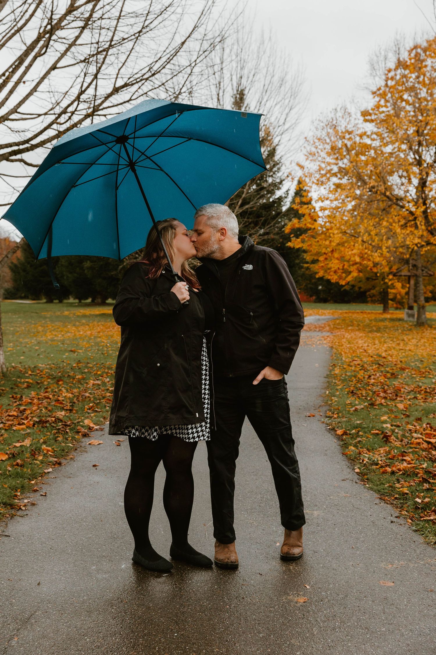 Two people share a romantic moment under a teal umbrella on a rainy autumn day with fallen leaves on the path.