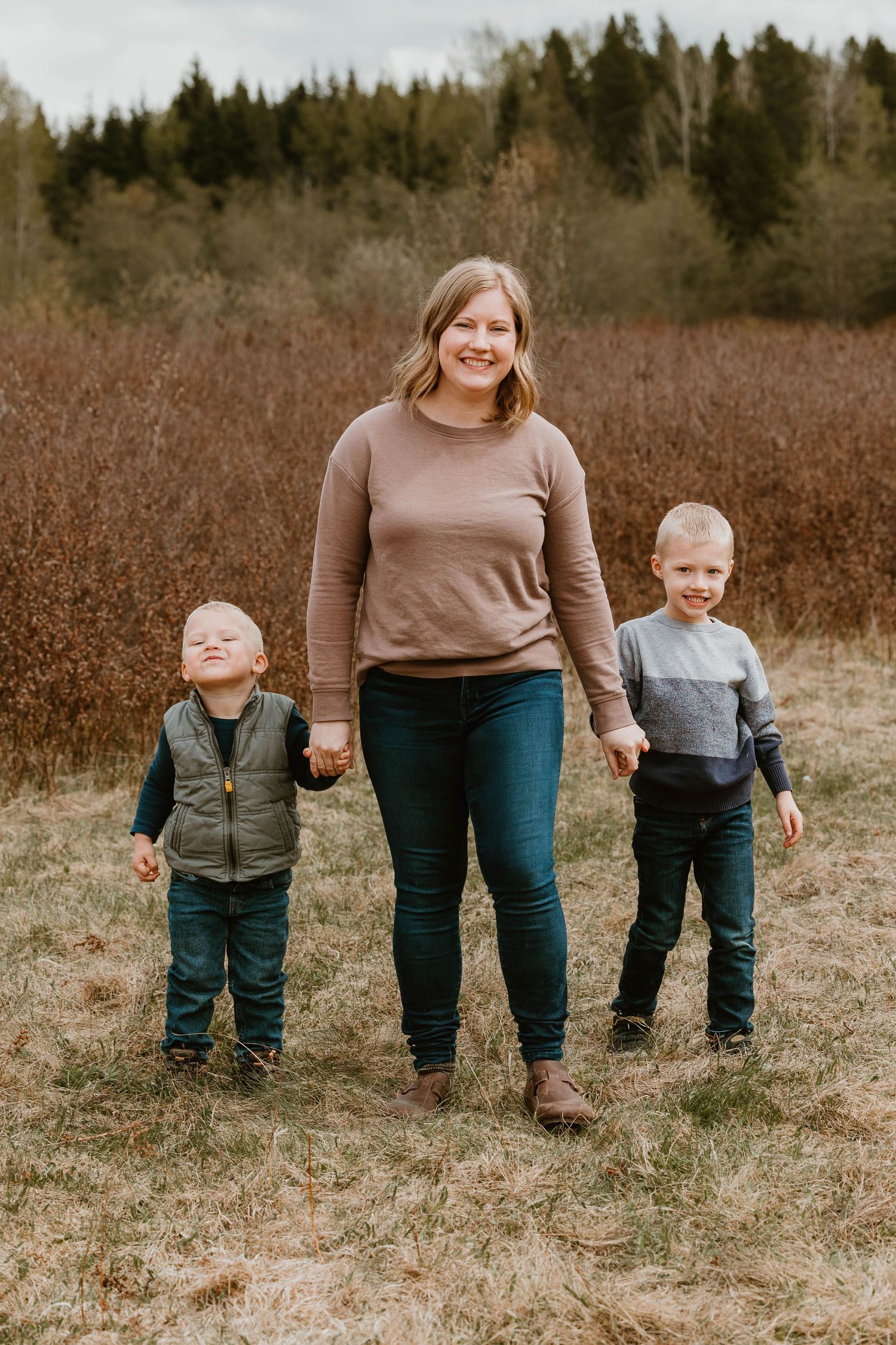A family group stands together outdoors in an autumn field with evergreen trees in the background.