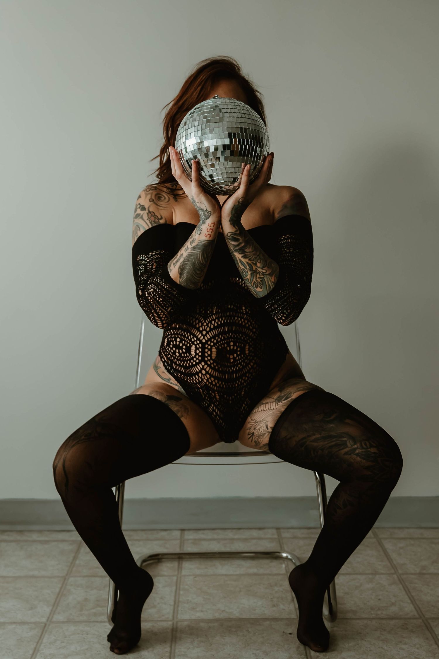 Figure in black lace bodysuit poses with a disco ball in an artistic boudoir photo.
