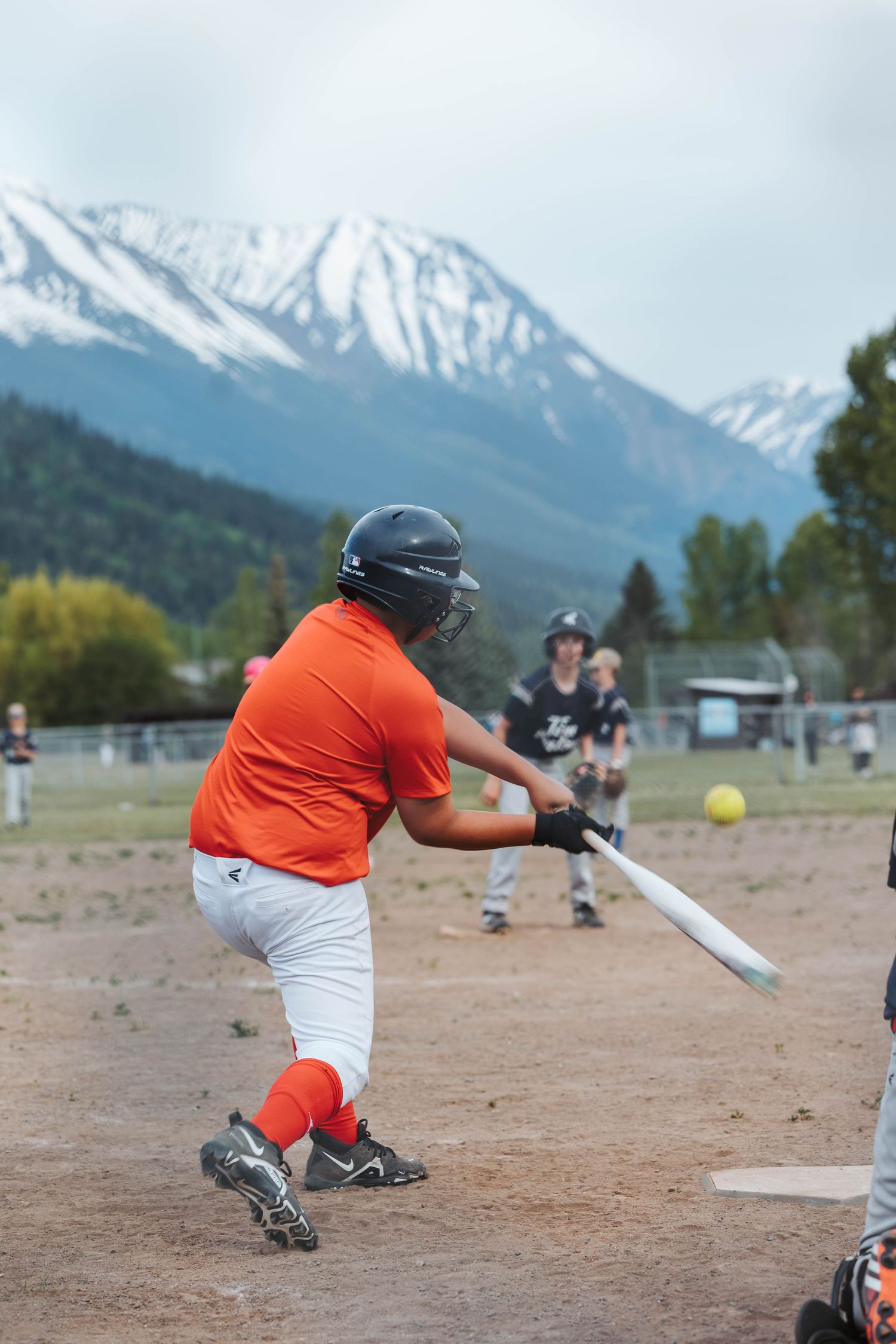 Baseball player in orange jersey swings at a pitch with snowy mountains in the background.
