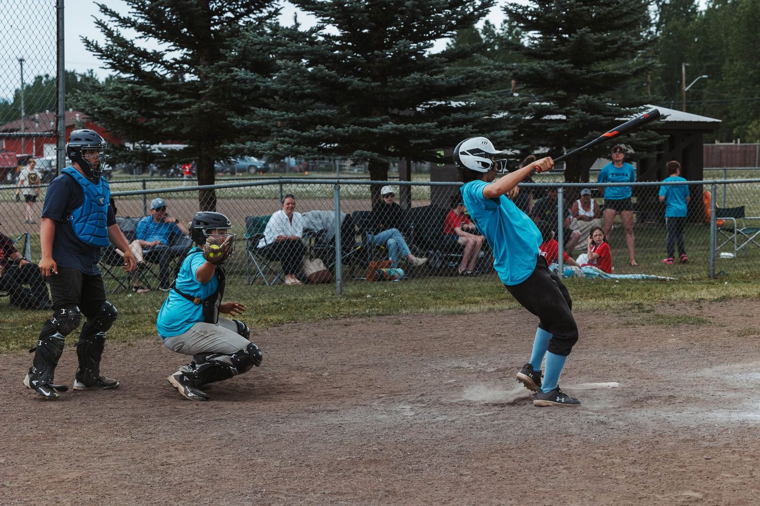 Youth baseball players in blue uniforms at bat and catching during a game on a dirt baseball field.