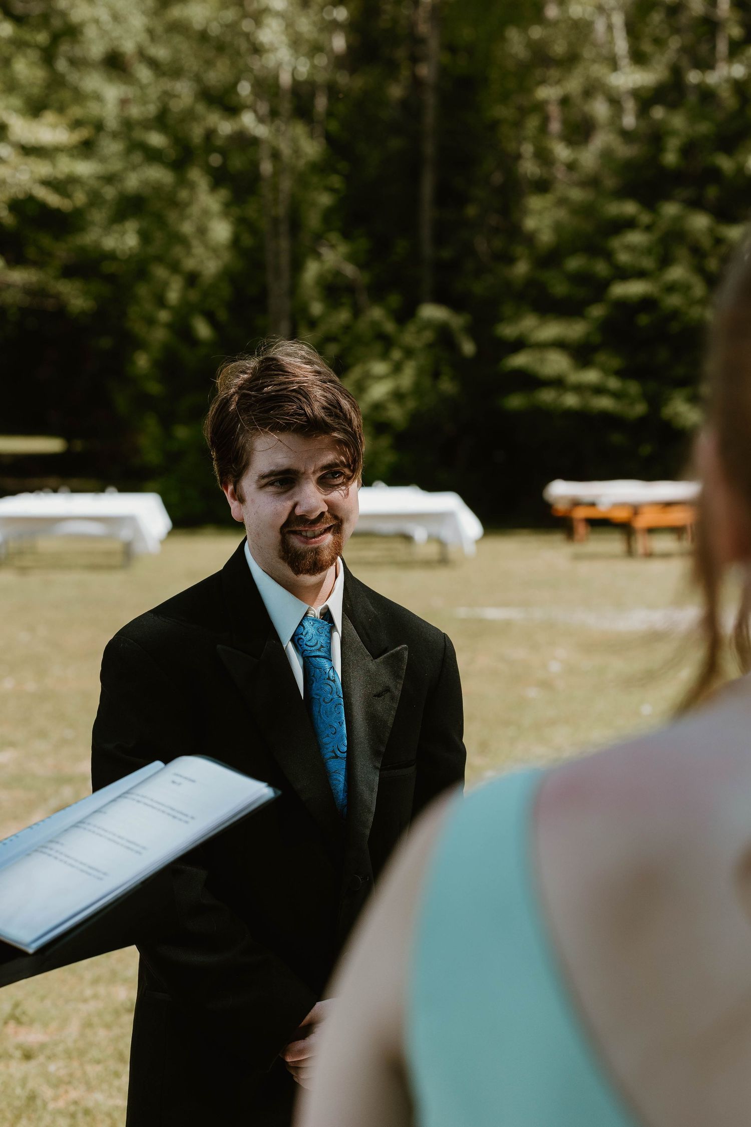 Groom in black suit and blue tie smiles during outdoor forest wedding ceremony.