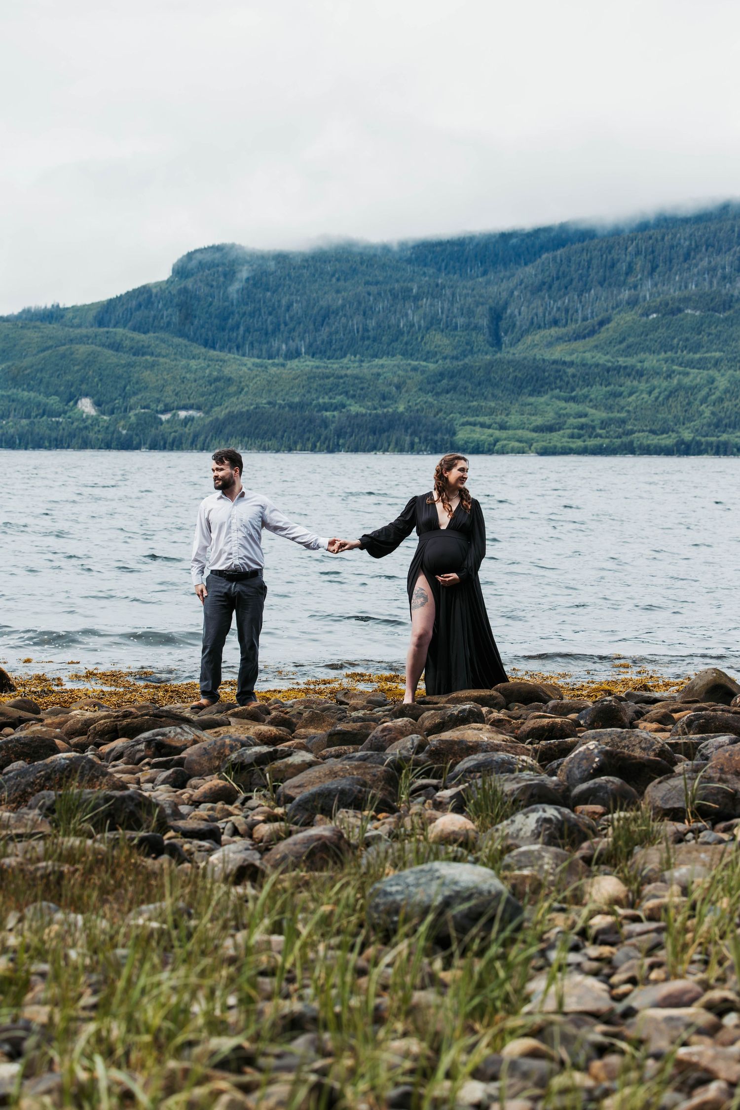 A couple poses on rocky shore with dramatic mountain landscape and ocean views.