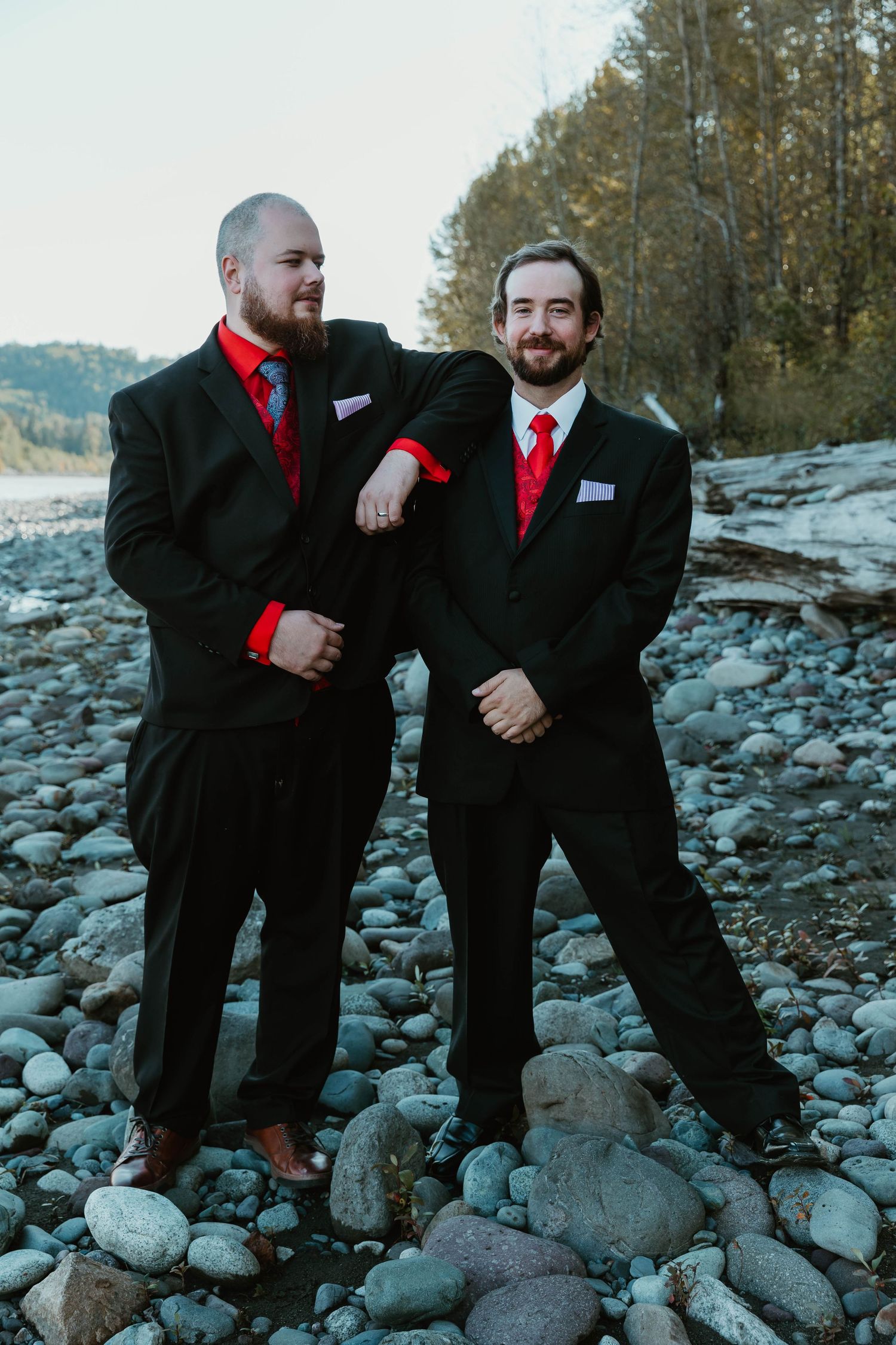 Two groomsmen in black suits with red ties pose on rocky riverbank.