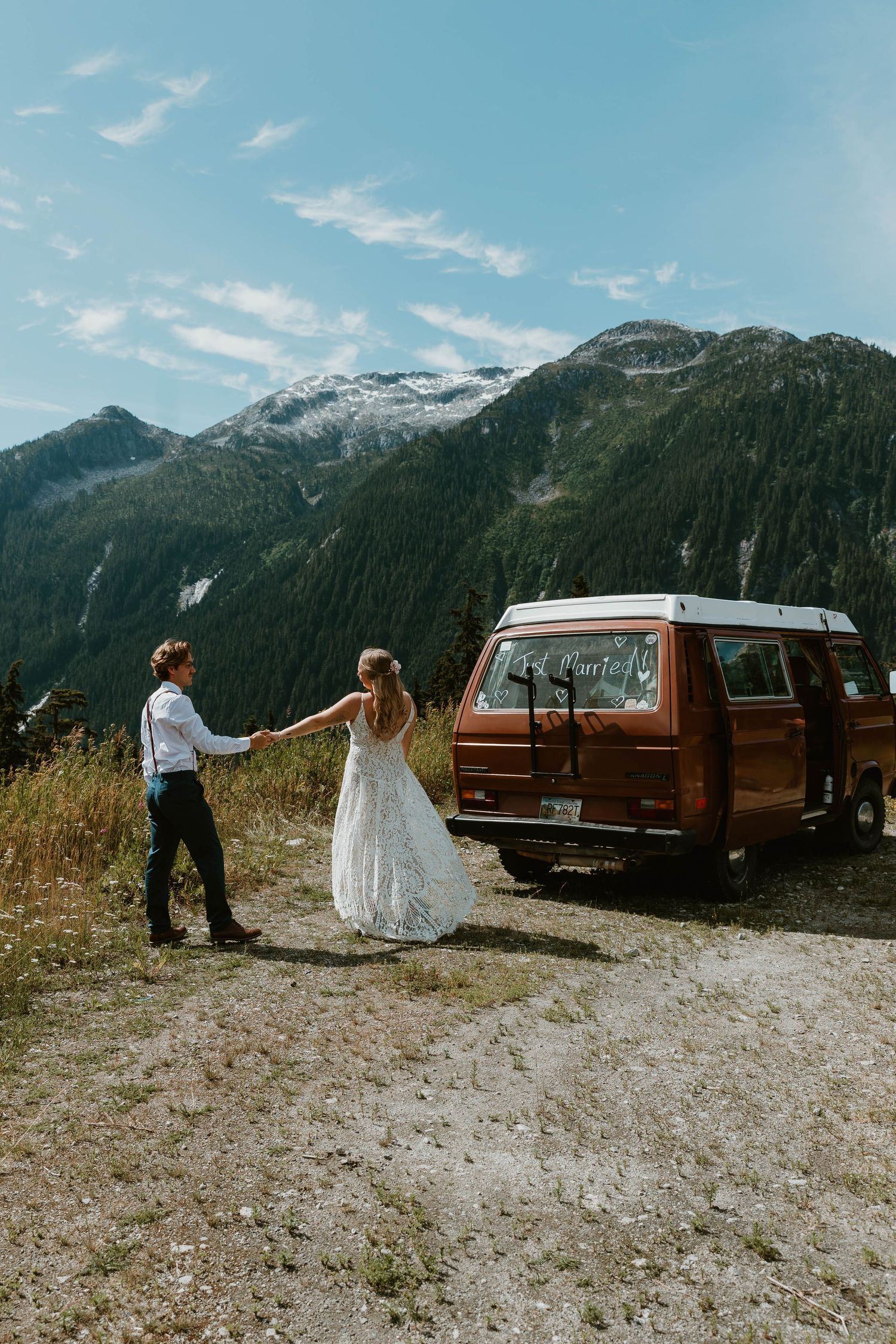 Couple poses by vintage brown van against dramatic mountain backdrop on their wedding day.