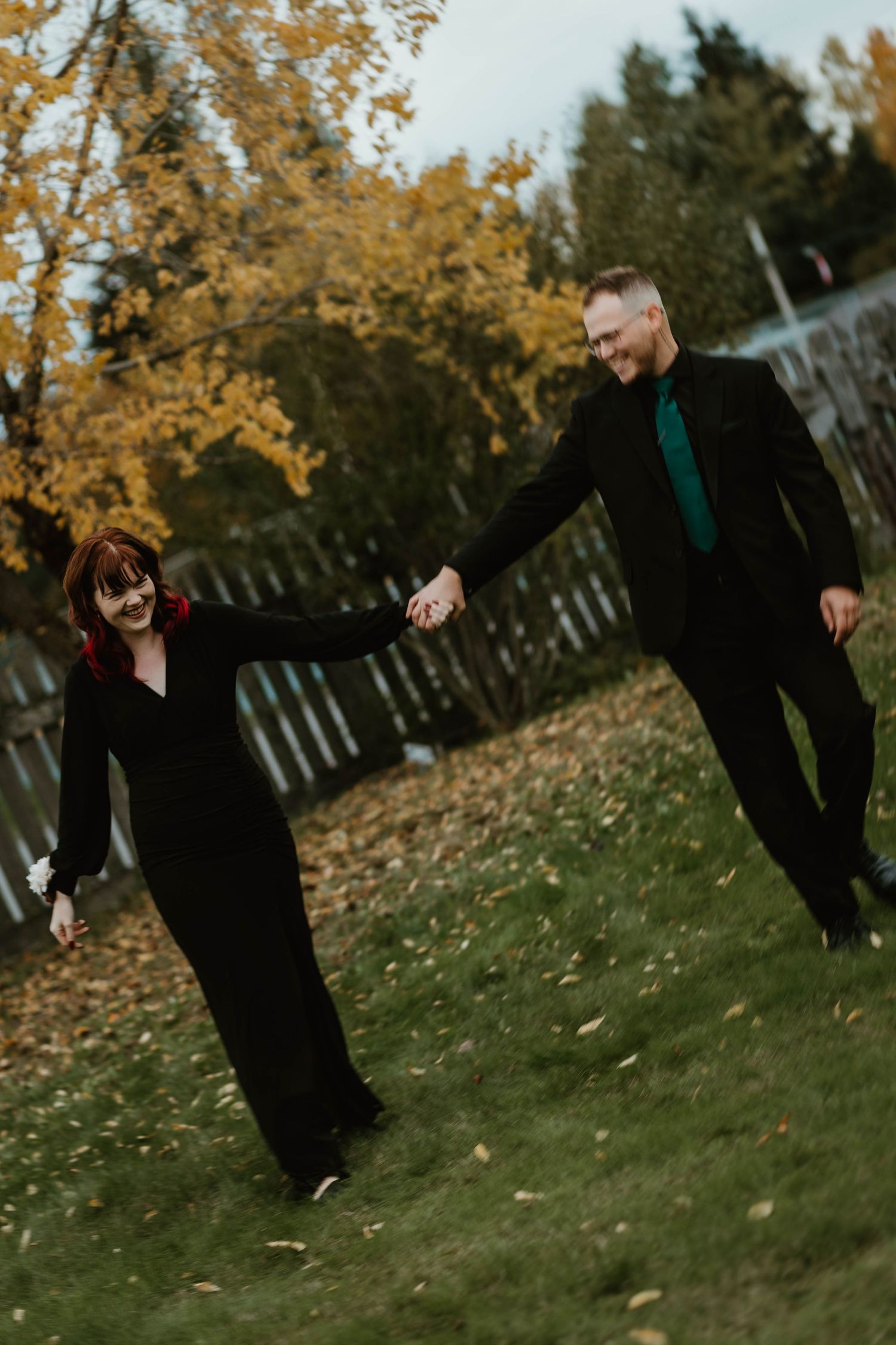 A couple holding hands while walking and laughing in a backyard with autumn leaves.