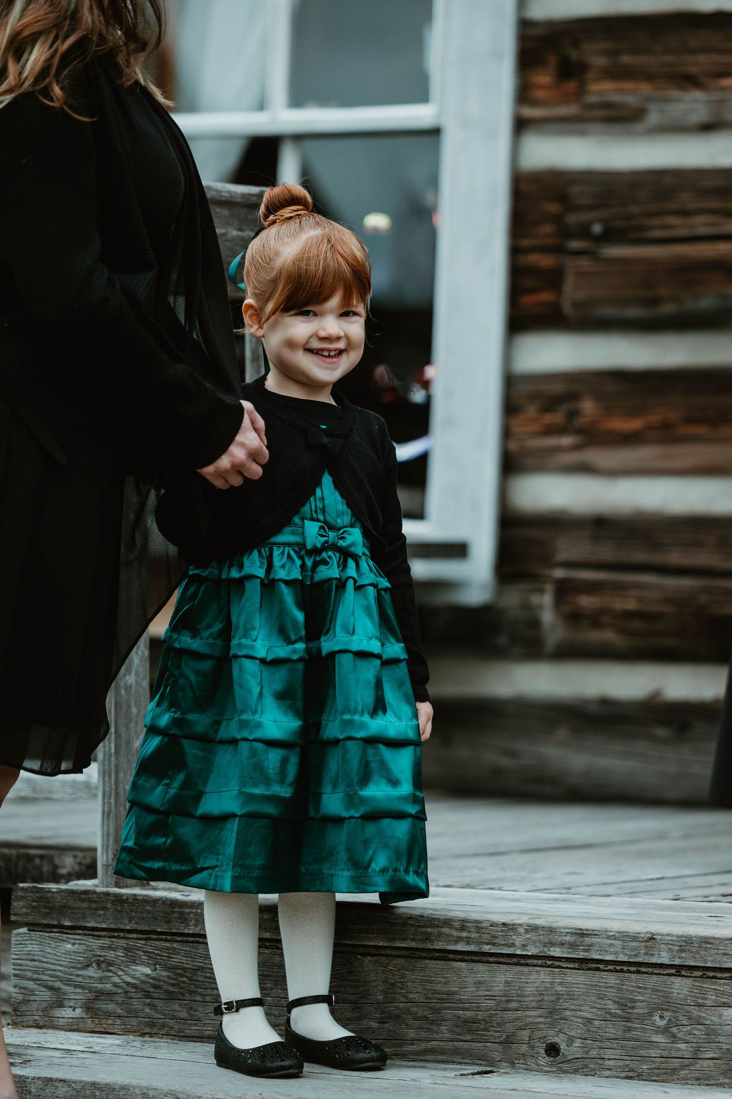 A small child wearing a teal ruffled dress stands on wooden steps by a rustic cabin.