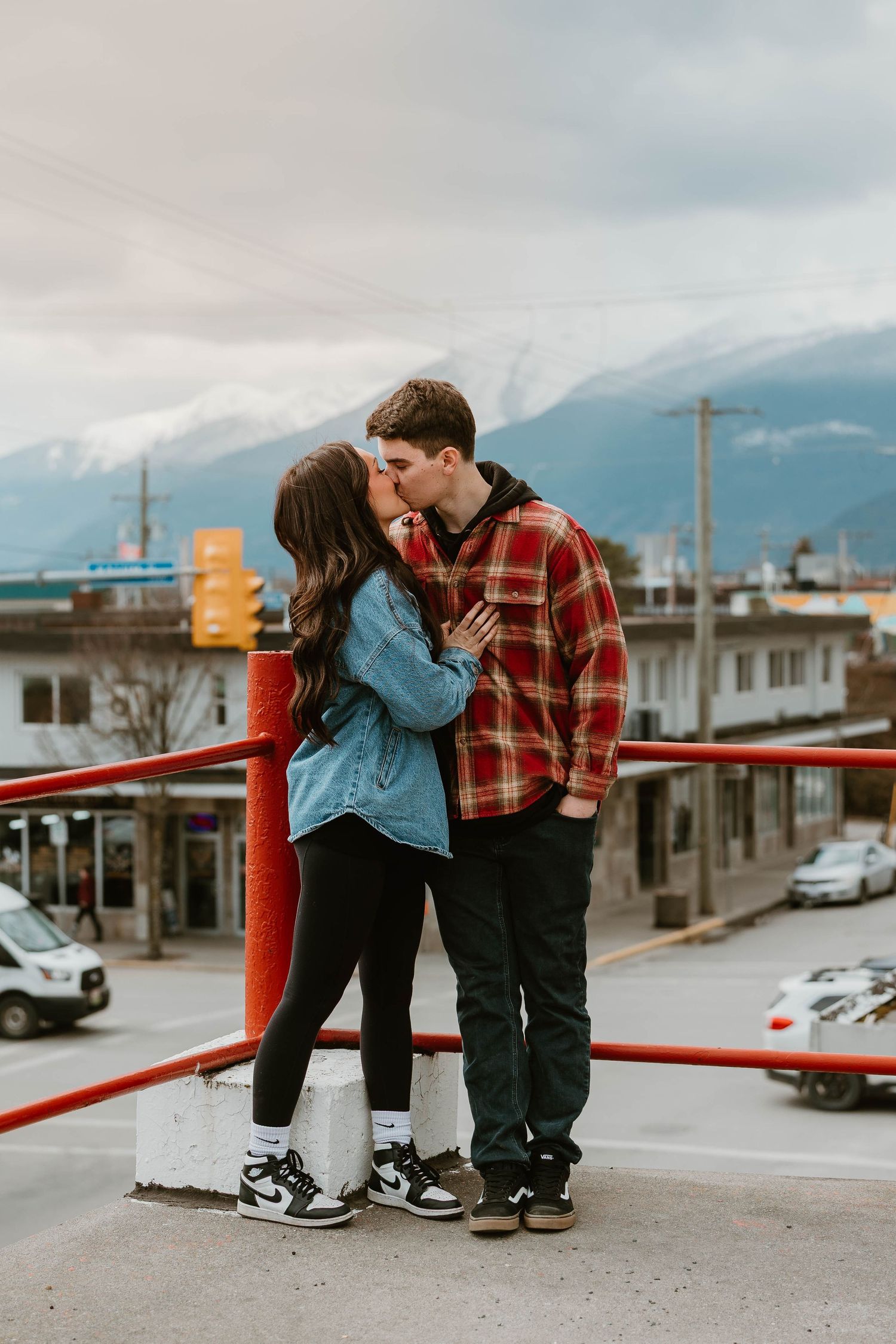 A couple shares a romantic moment in denim and plaid against a mountain backdrop in an urban setting, standing close together.